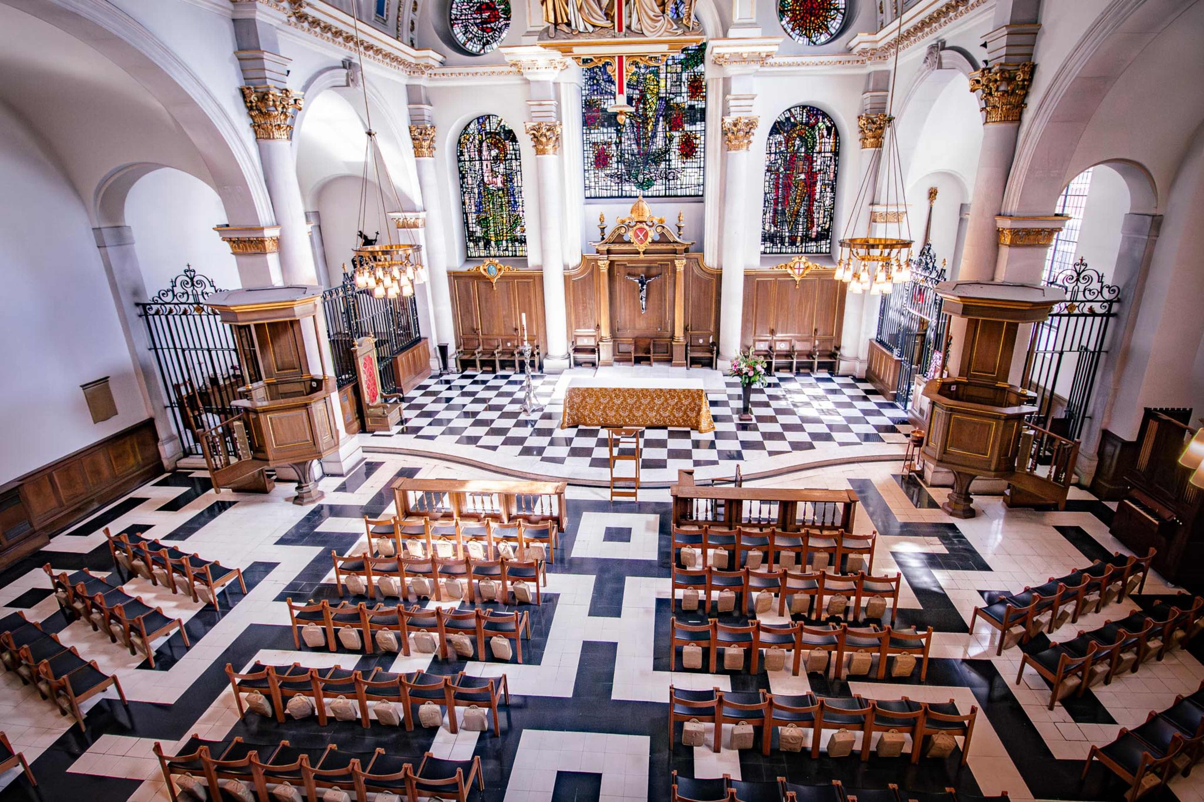 The image shows the interior of a church featuring a symmetrical layout with a checkered floor, an altar at the front, and rows of wooden chairs facing the altar.