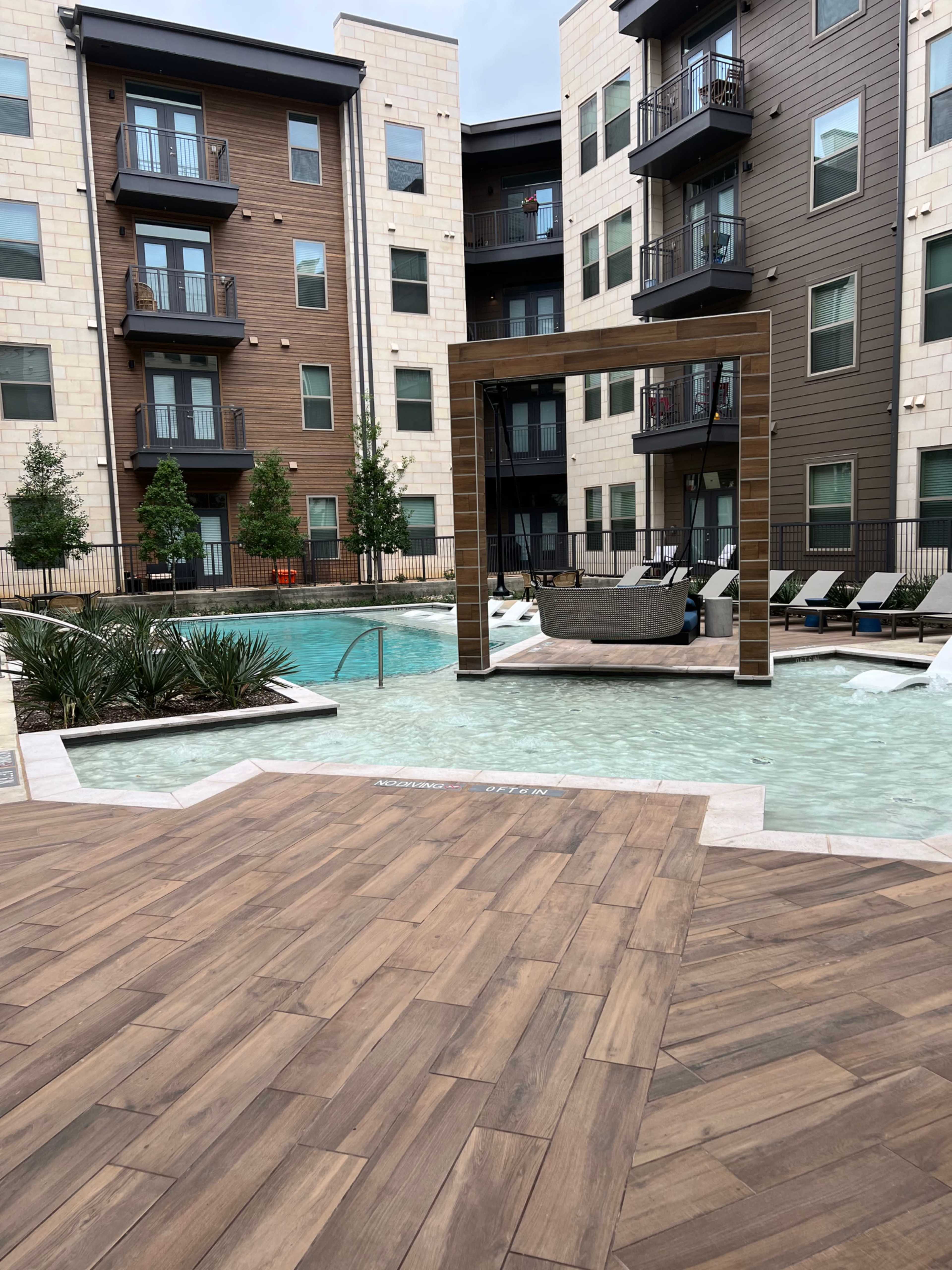 A modern apartment complex with a pool area featuring a wooden pergola, surrounding lounge chairs, and landscaped greenery.