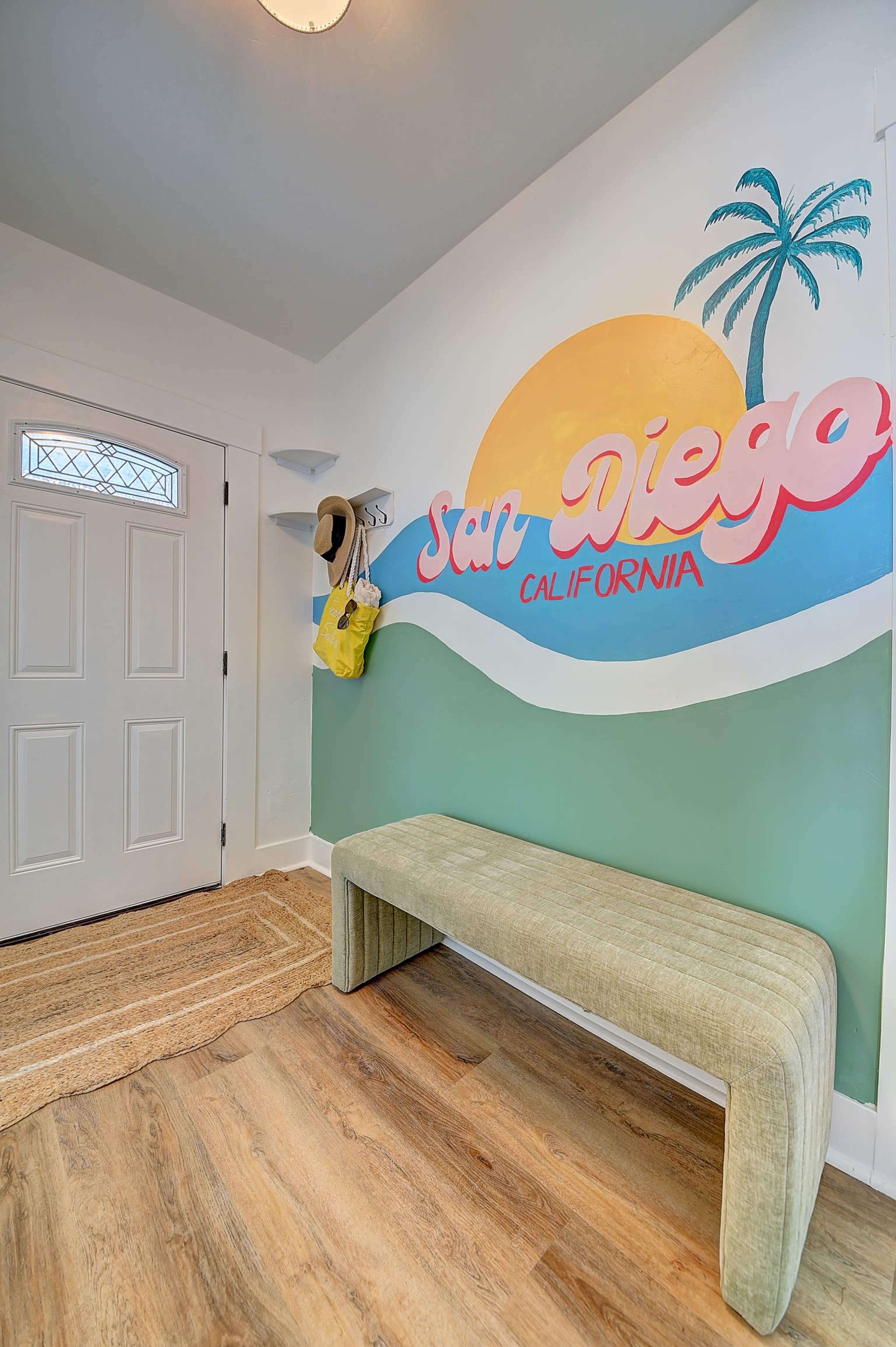 The entryway features a green wall with a colorful mural that reads "San Diego California," a bench, and a door, alongside a hat and bag hanging nearby.