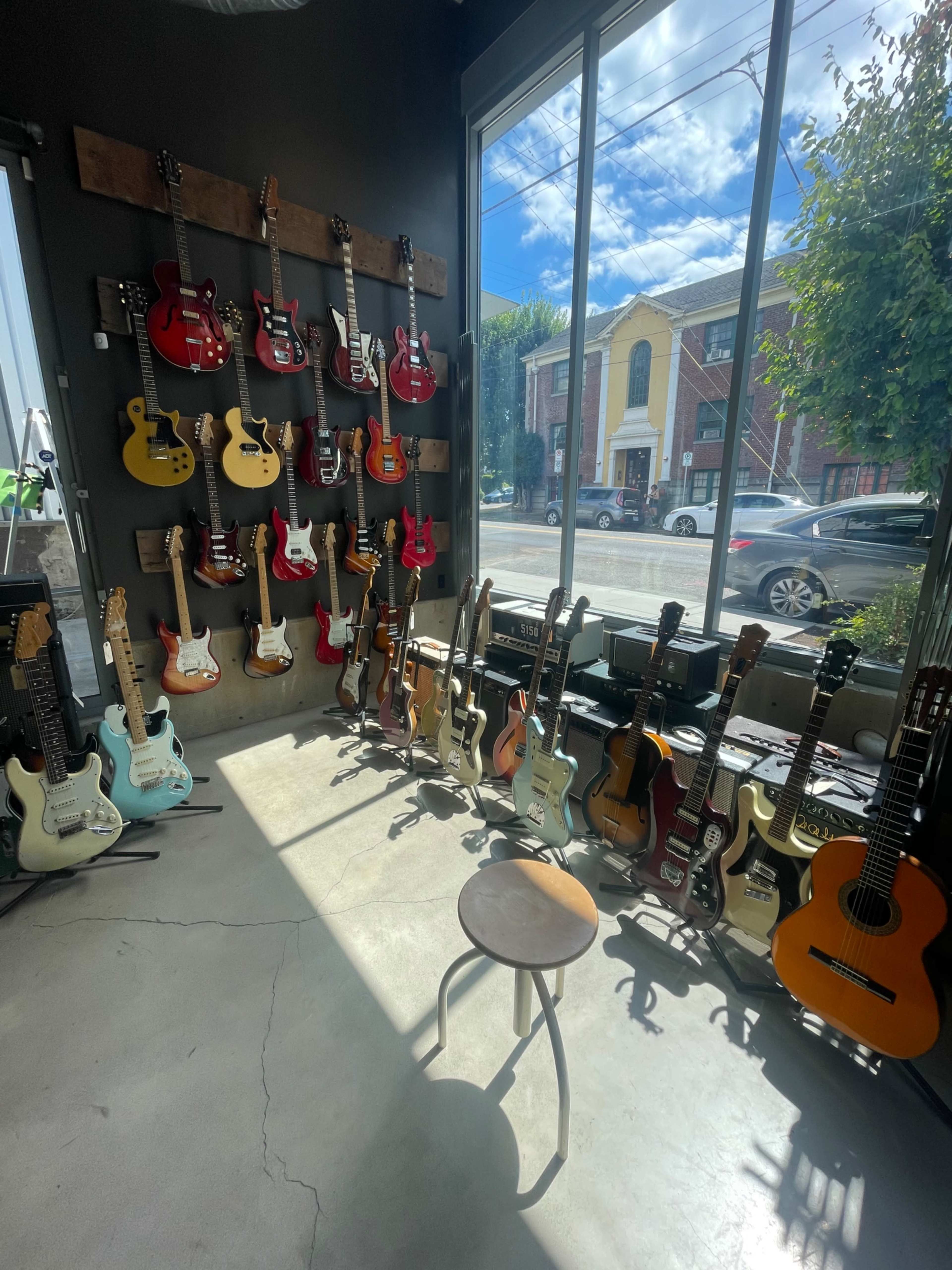 A guitar store with a wall of various electric guitars and several acoustic guitars arranged on the floor near a stool, with sunlight streaming through large windows.