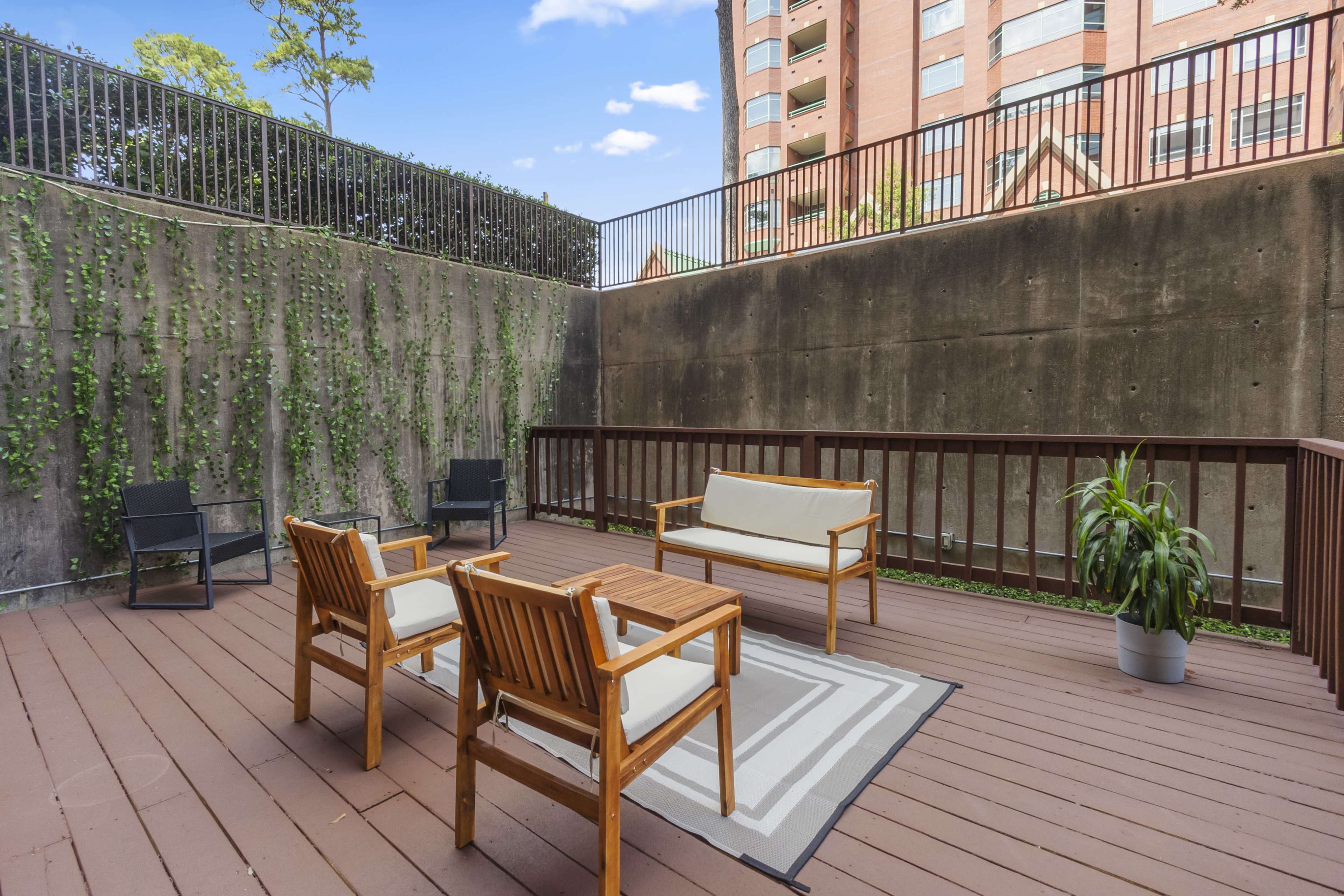 The image shows a wooden deck area with seating consisting of three chairs and a coffee table, bordered by a concrete wall and greenery.
