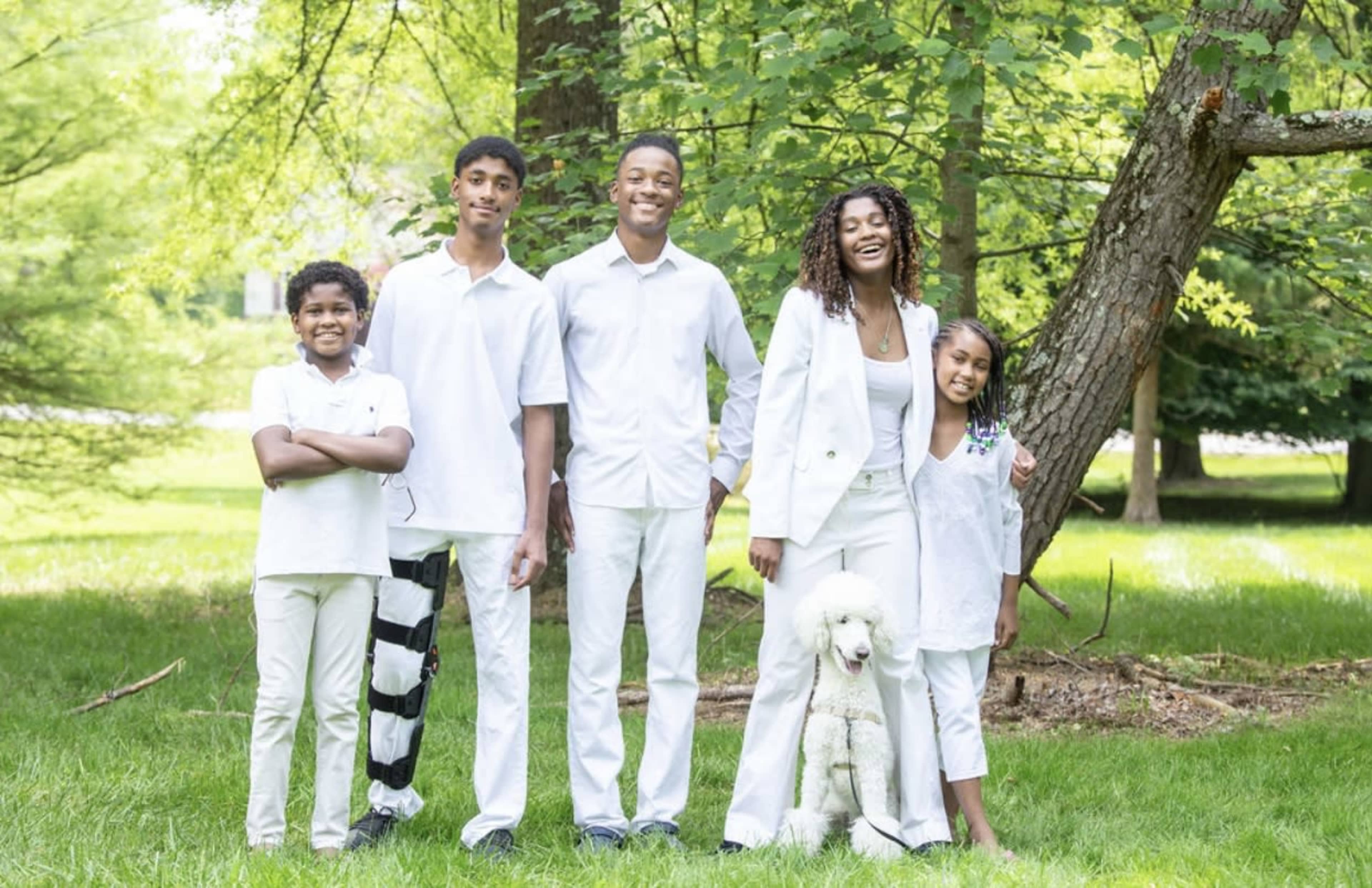 A family of five, dressed in white, stands together in a grassy area surrounded by trees, with one member holding a poodle.