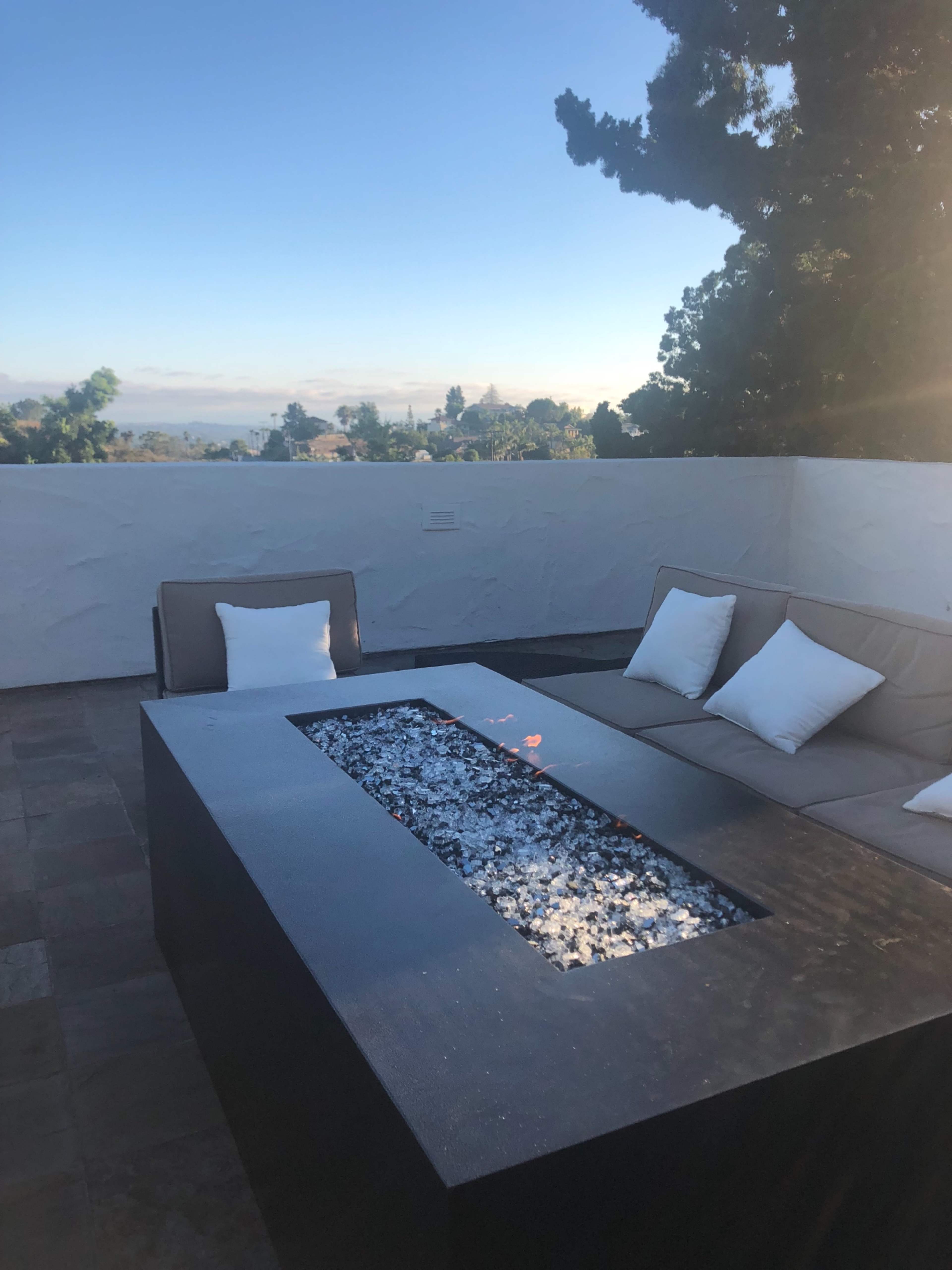 A modern outdoor seating area features a fire pit with a glass stone surface and a sectional sofa with white cushions, set against a clear sky and distant landscape.