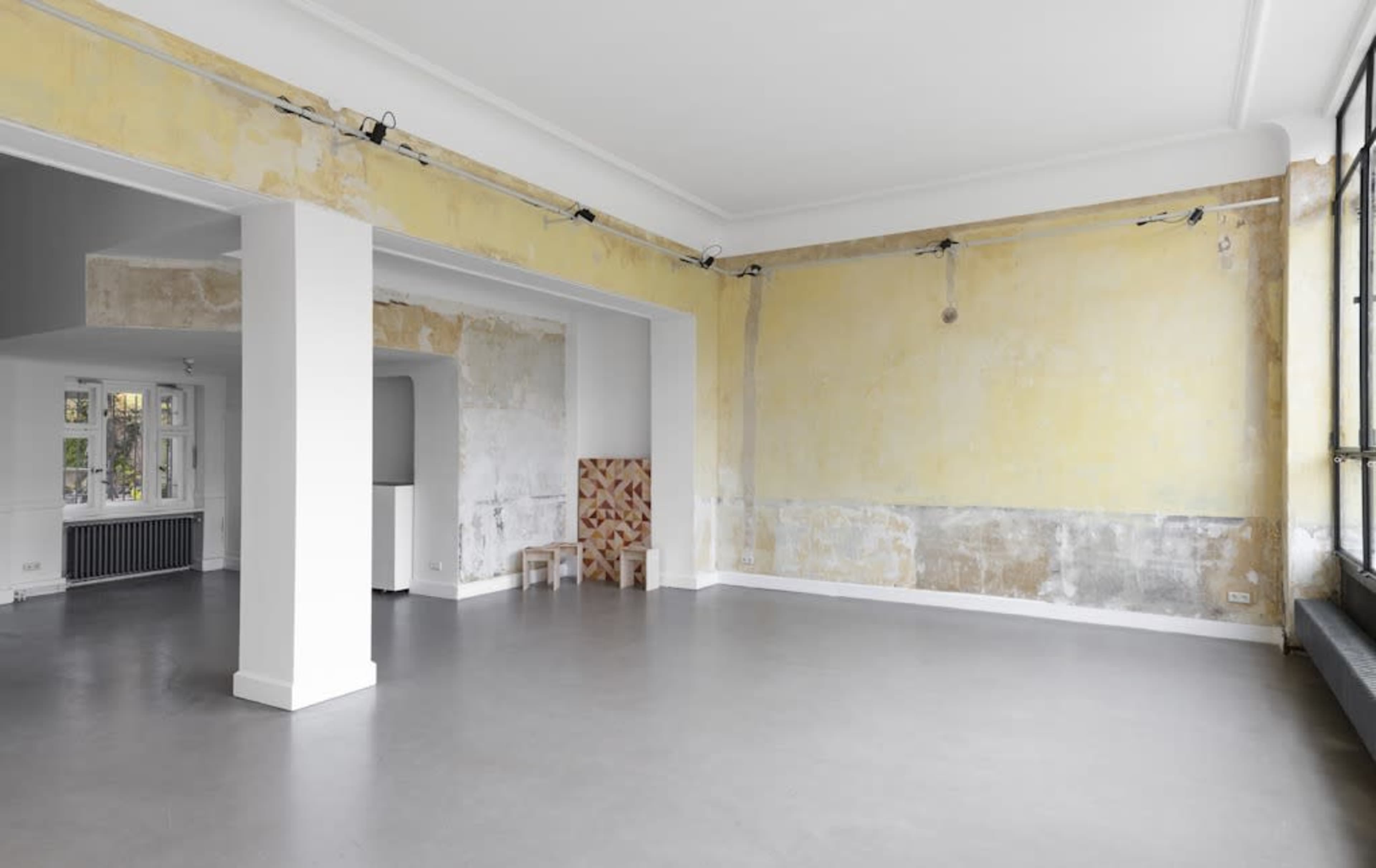 The image shows a spacious room with bare walls that have a faded yellow color, polished concrete floors, and minimal furnishings.