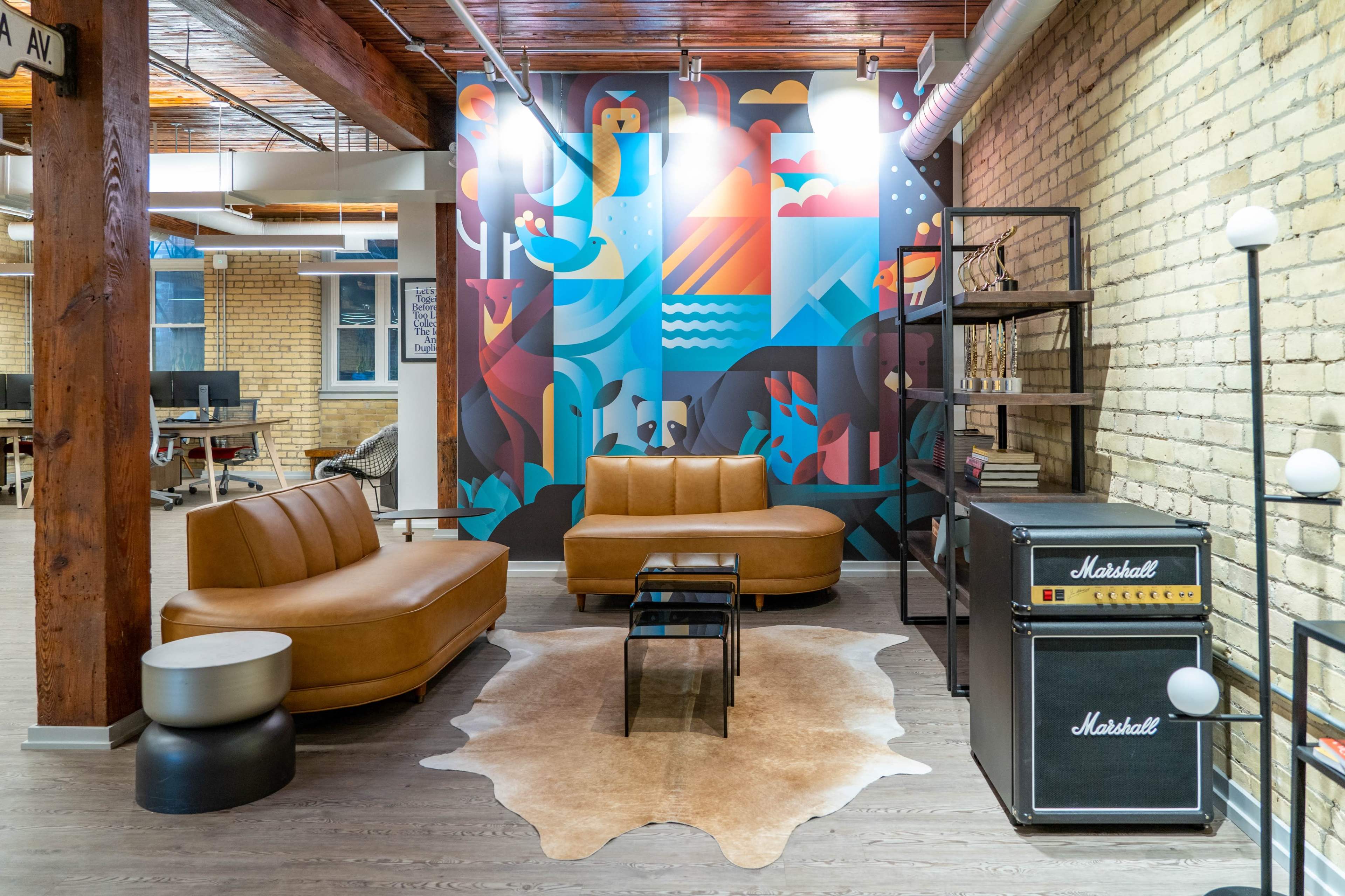 A modern office lounge area featuring a brown leather sofa, a coffee table, a decorative rug, and a black Marshall speaker cabinet against a colorful mural on the wall.
