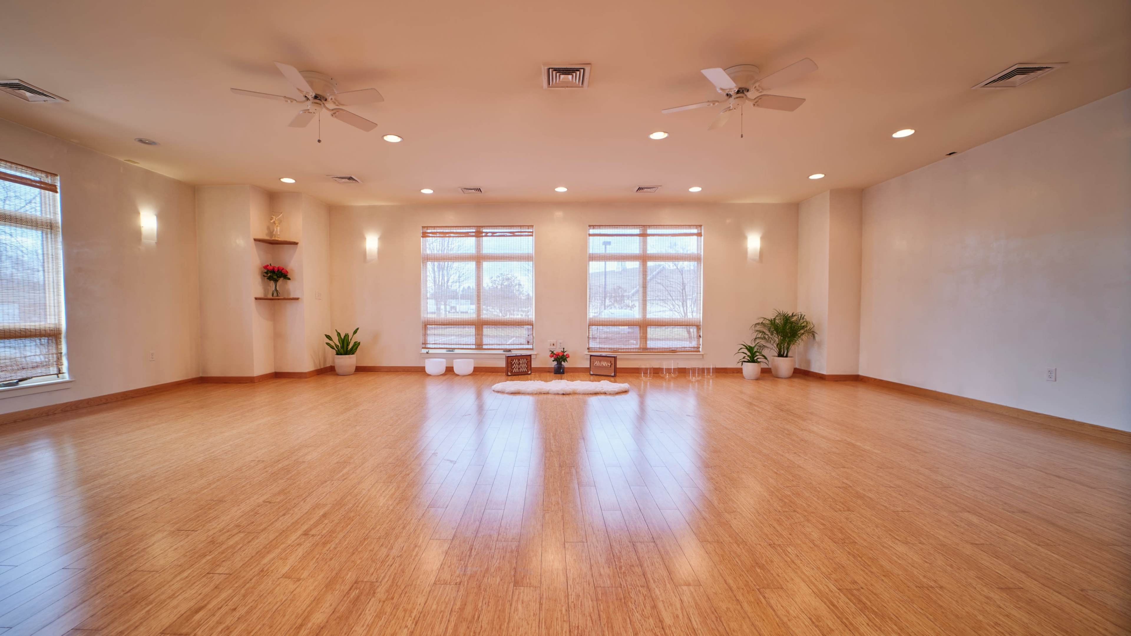A spacious yoga studio with bamboo flooring, large windows, and several plants arranged around the room.