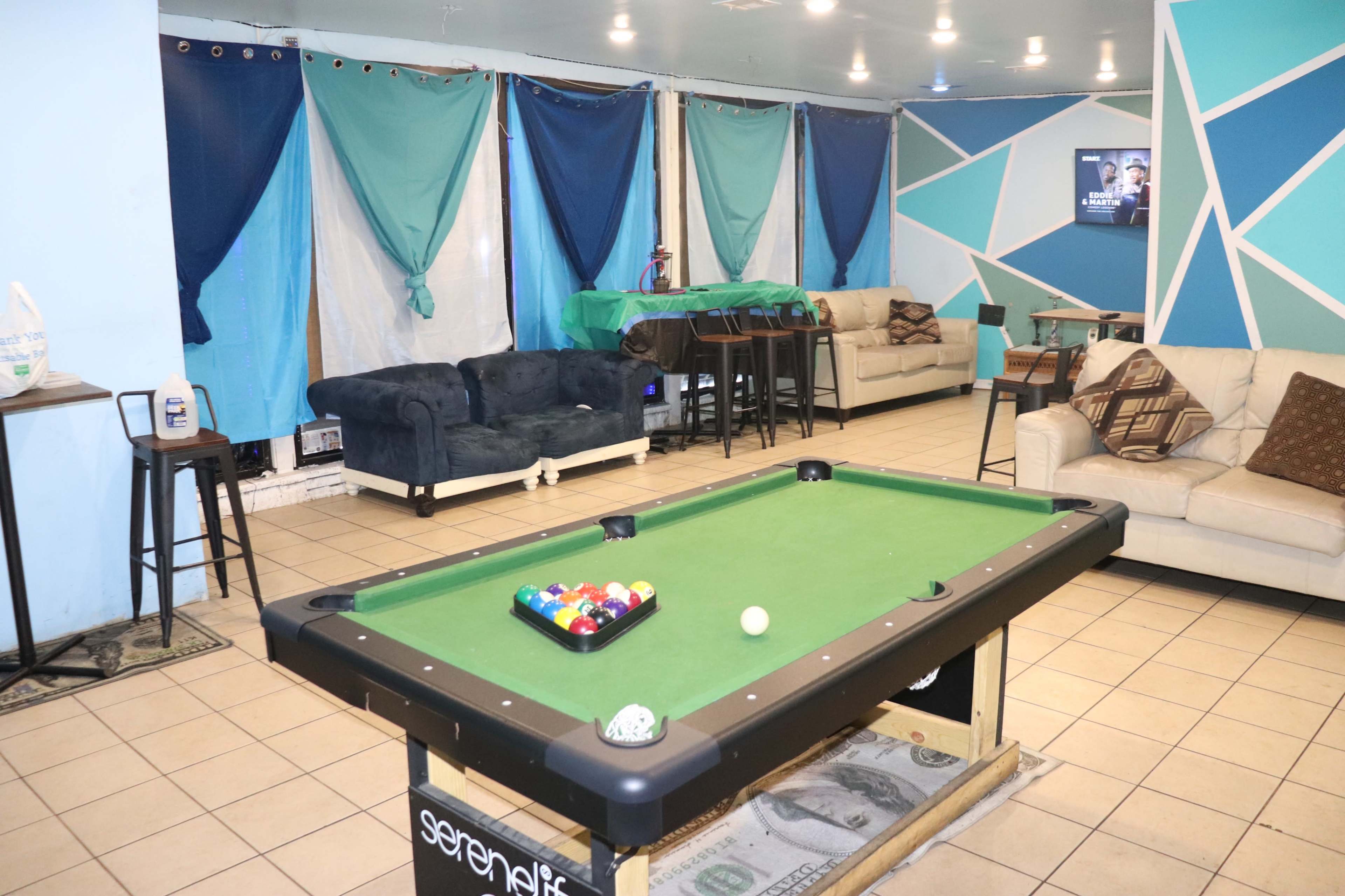A recreation room featuring a green pool table with balls, two couches, and a table in front, all set against a backdrop of blue and teal drapes.