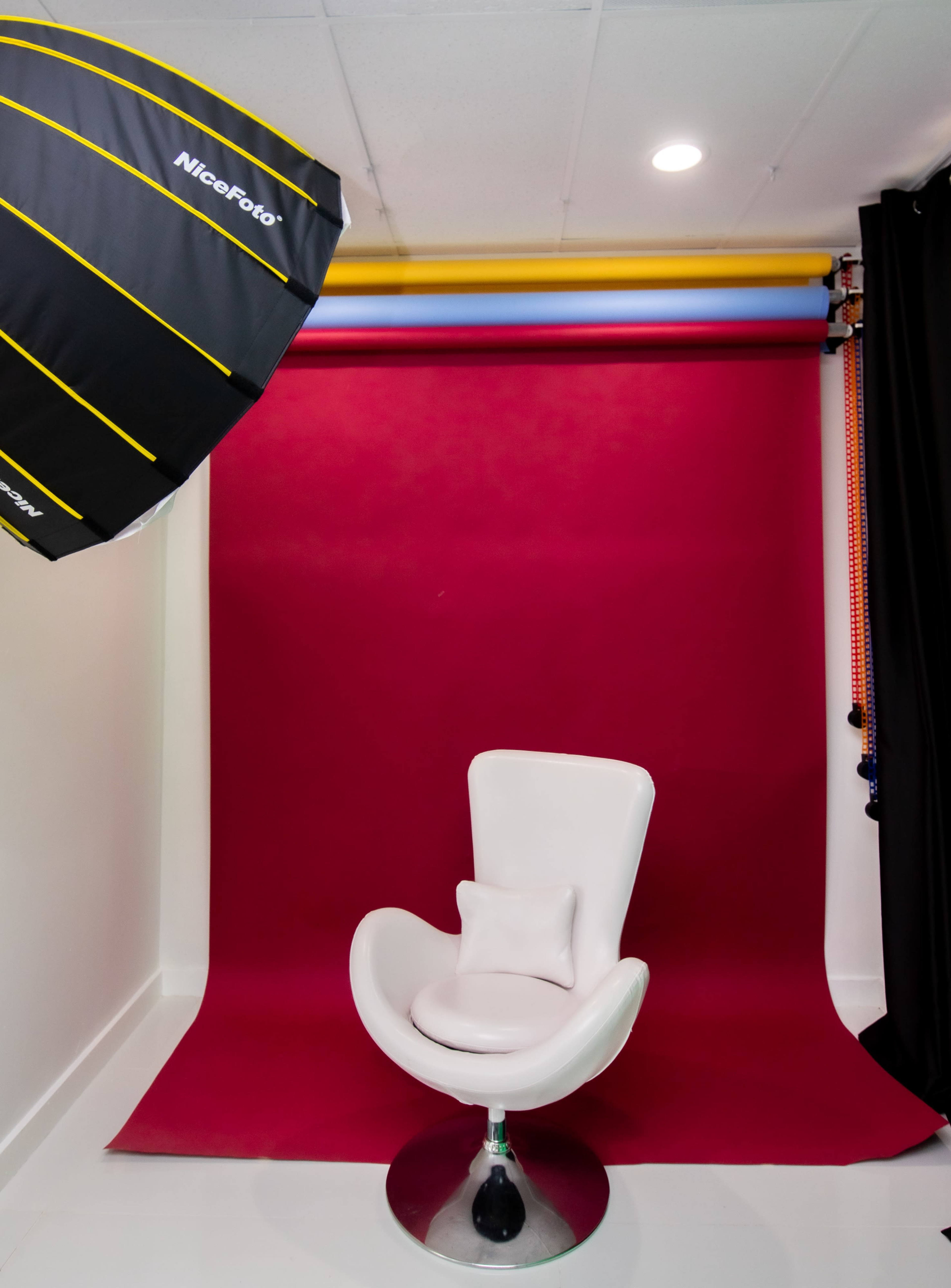 A photography studio setup with a white chair positioned in front of a burgundy backdrop, accompanied by a large softbox lighting overhead.