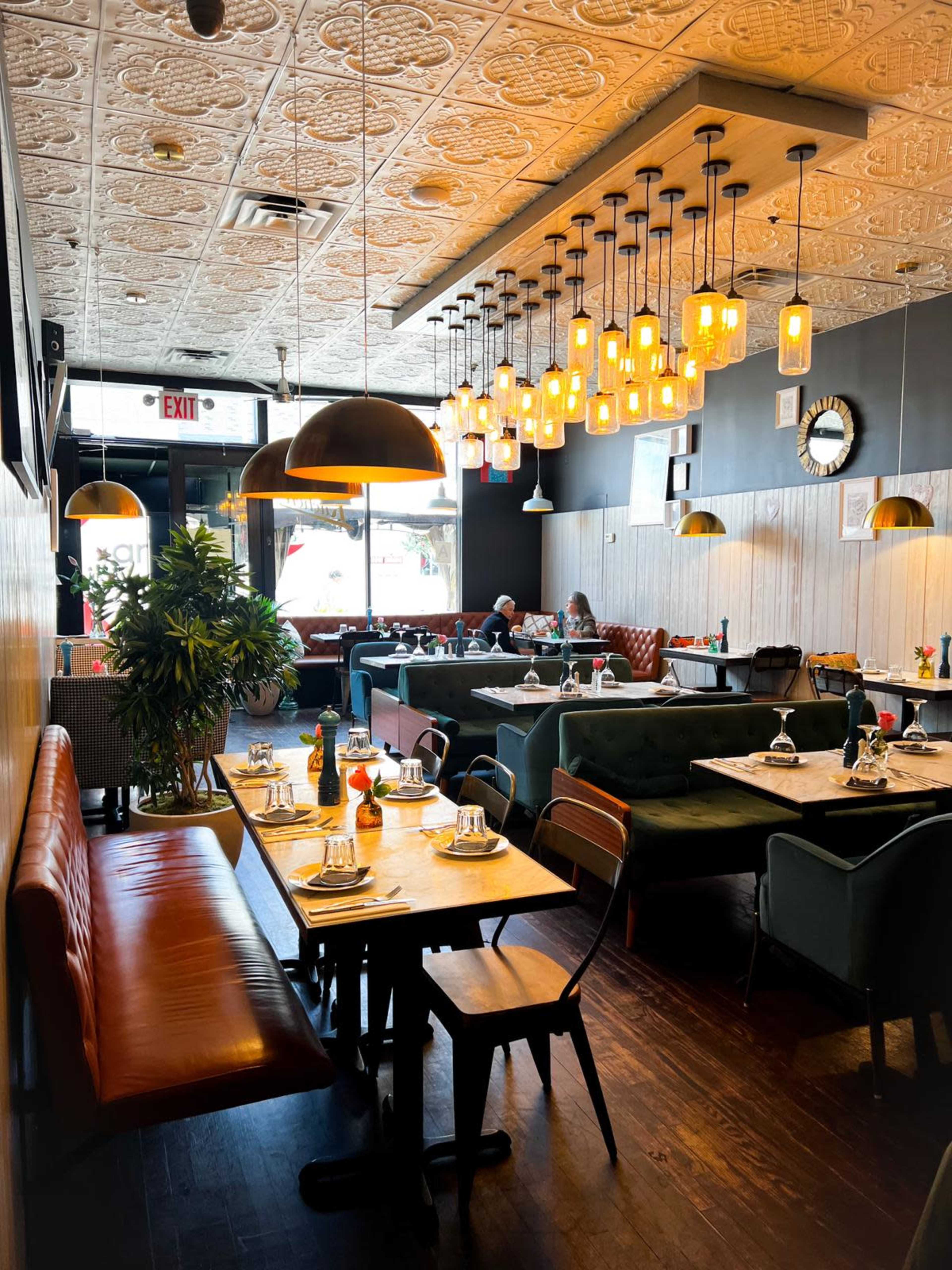 The interior of a restaurant features wooden tables and a variety of seating options, illuminated by pendant lights, with a view of patrons dining by the windows.