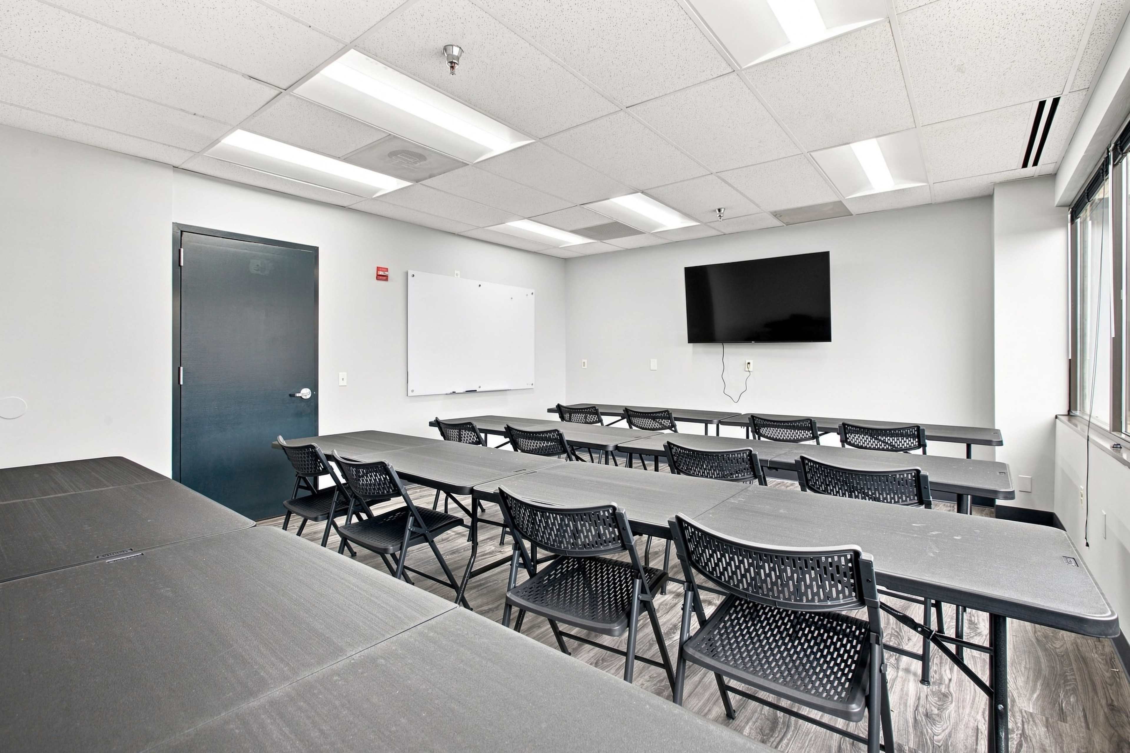The image shows a classroom with several rows of black folding chairs and tables, a whiteboard, and a wall-mounted television.
