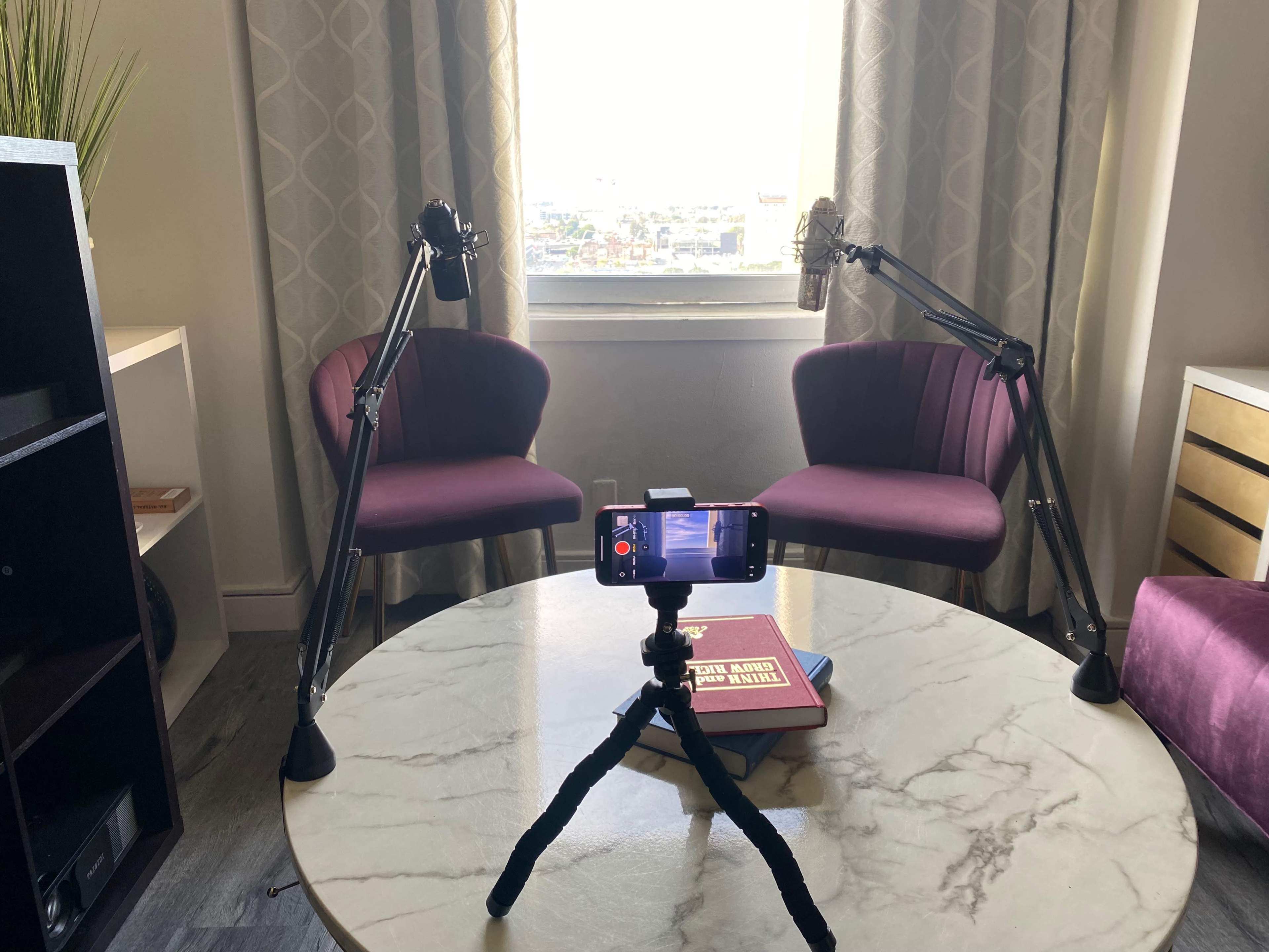 A setup for a podcast with two microphones attached to adjustable arms facing two purple chairs and a smartphone on a tripod recording on a marble table.