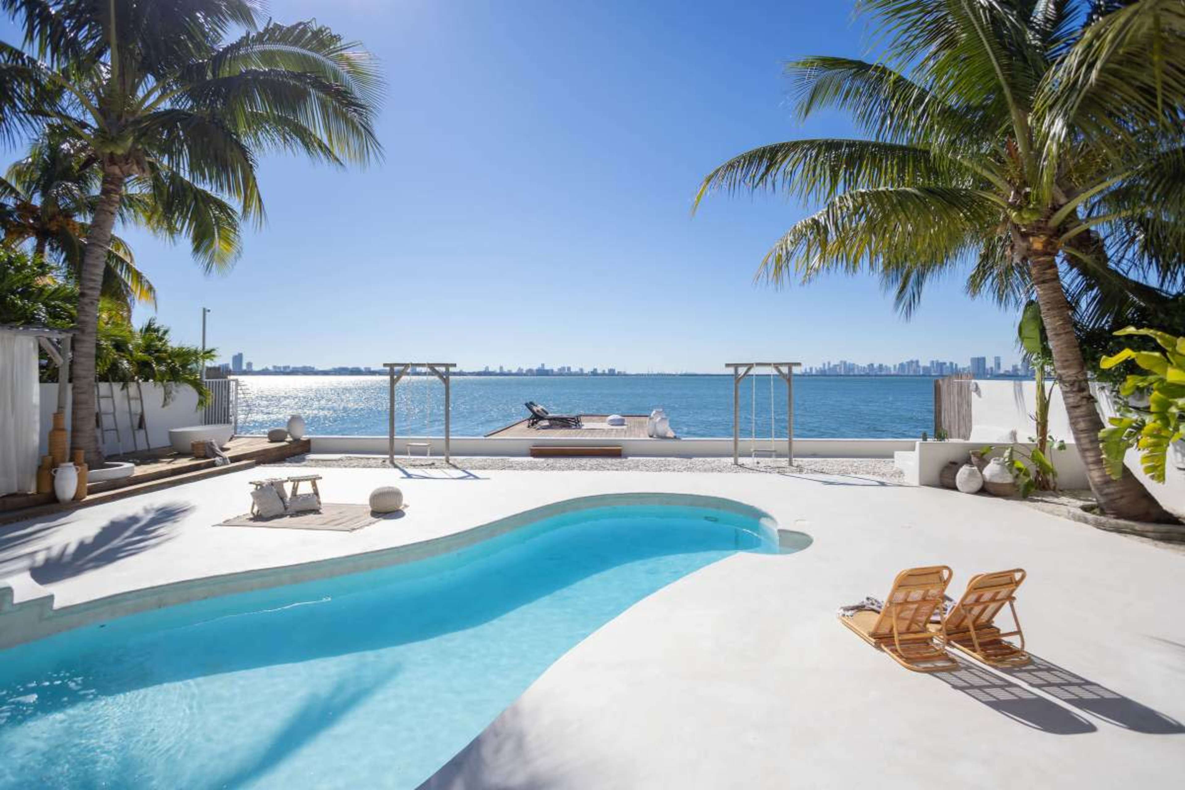 The image shows a curved swimming pool in the foreground with lounge chairs beside it, overlooking a tranquil sea and a skyline in the background under a clear blue sky.