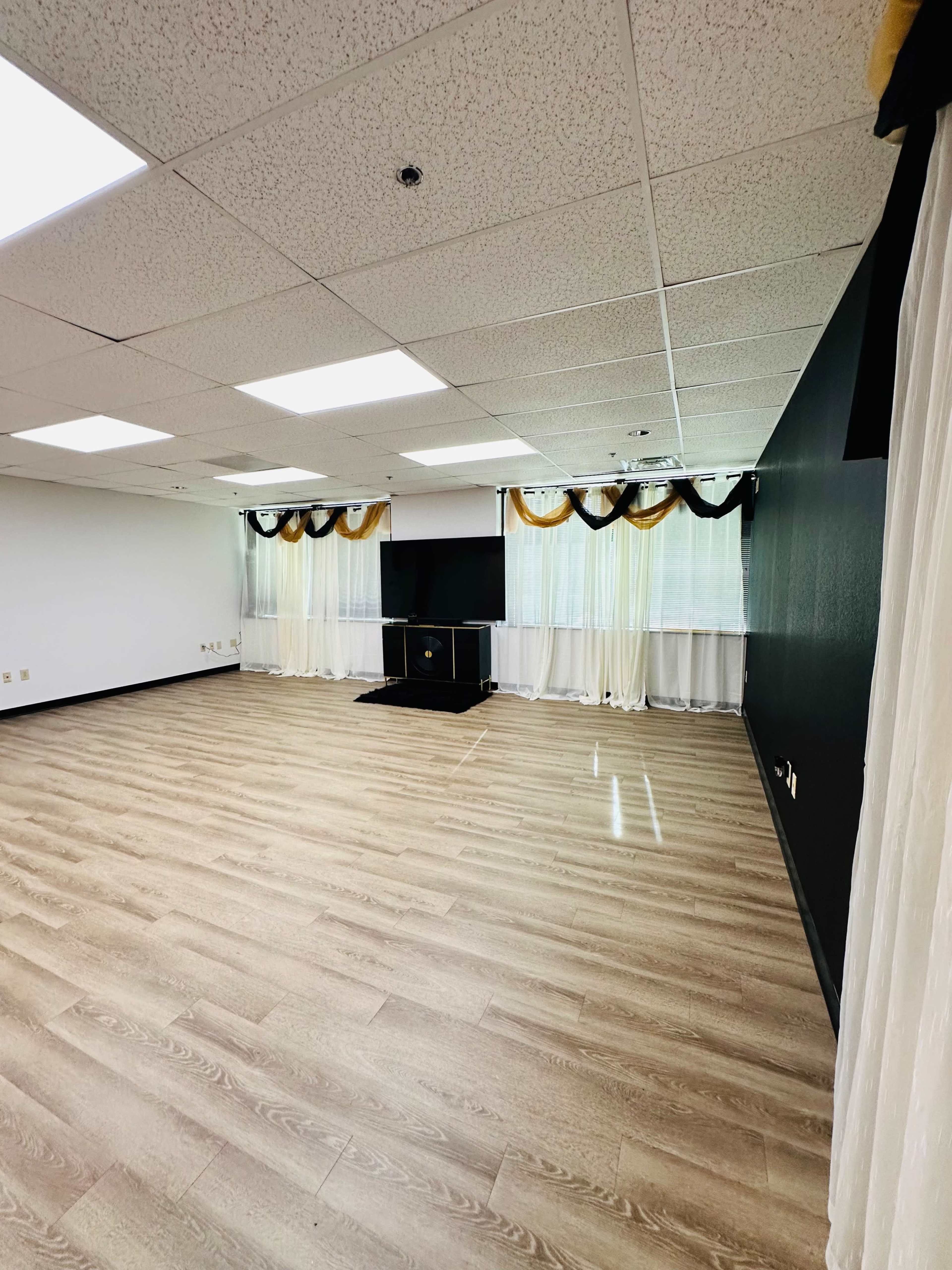 The image shows a spacious, empty room with wooden flooring, a television set on a stand, and curtains with decorative ties at the windows.