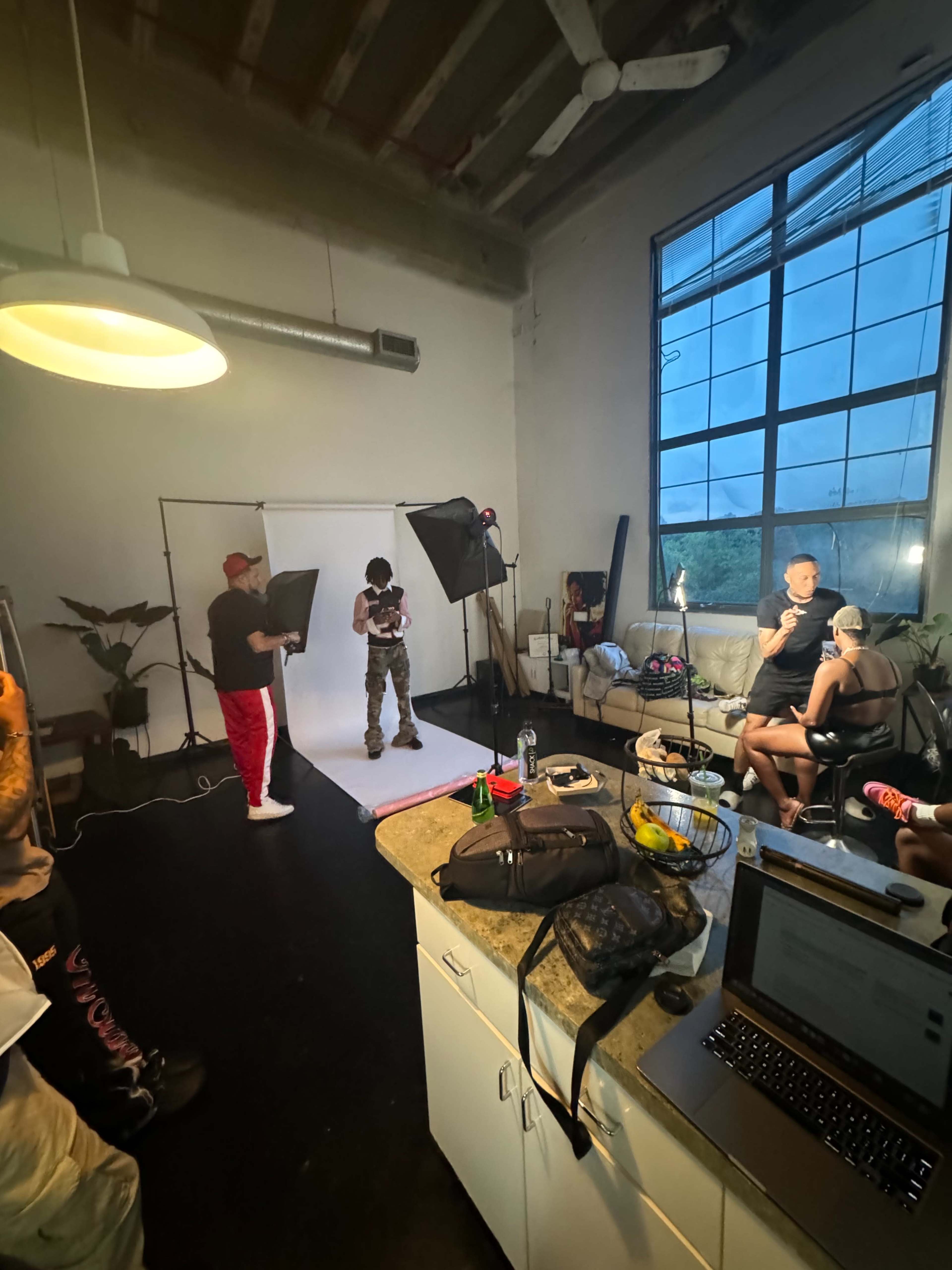 The image shows a photography studio with a model posing in front of a backdrop, surrounded by crew members and equipment, while some people are seated nearby.