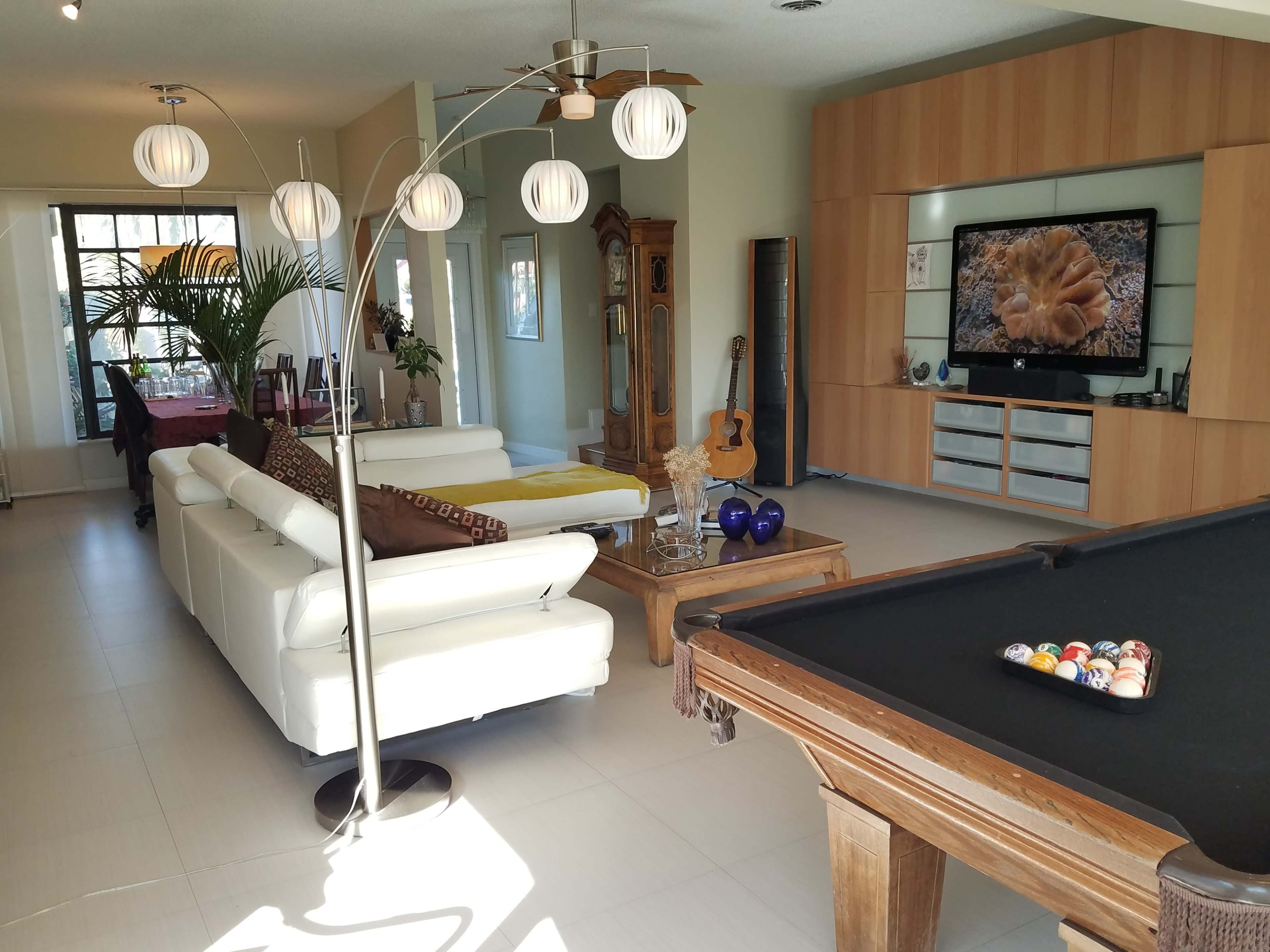 The image shows a spacious living area featuring a white sectional sofa, a wooden pool table, a large television mounted on a wall unit, and decorative plants.