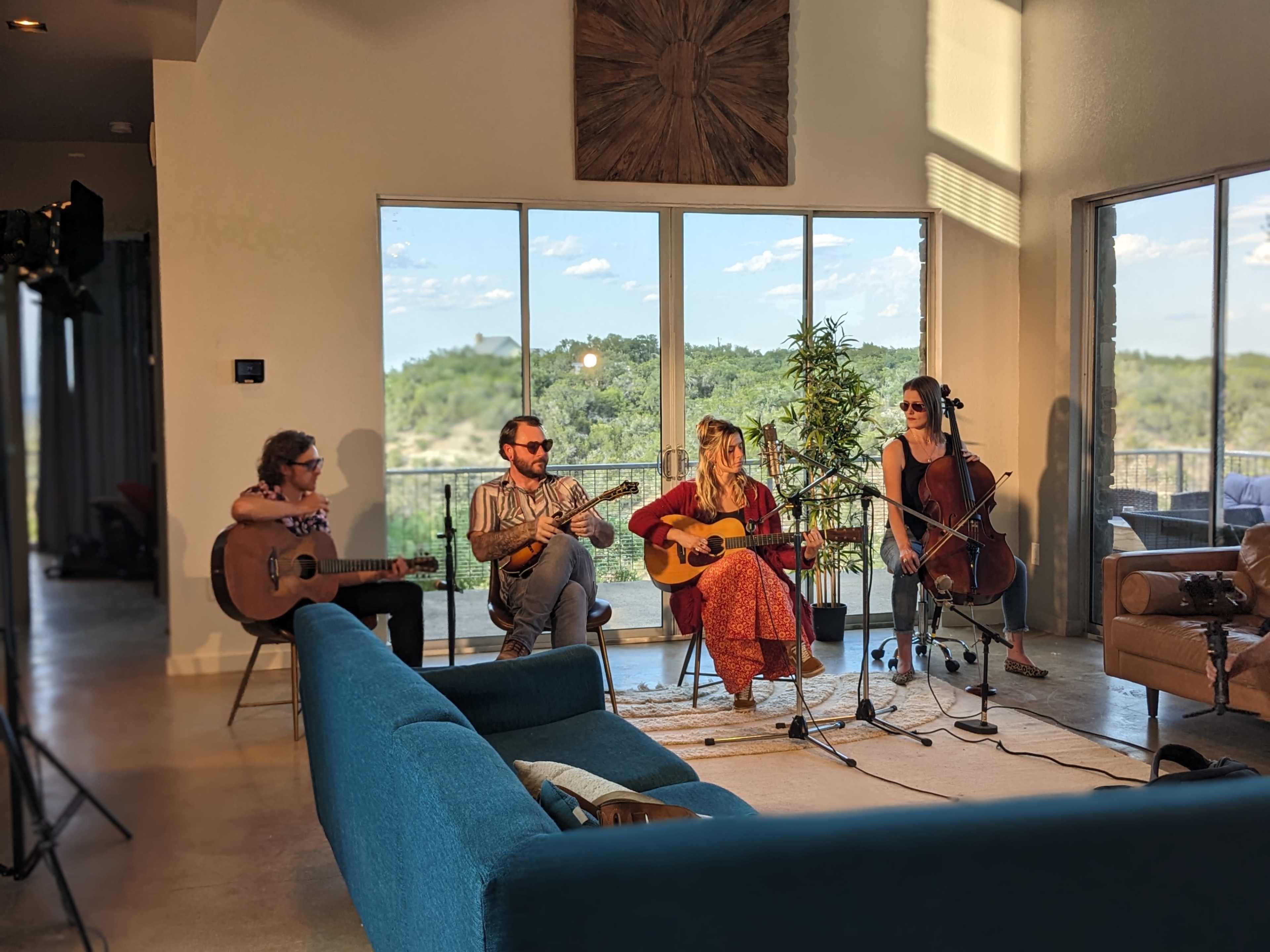 Four musicians play instruments in a bright living room with large windows overlooking a green landscape.