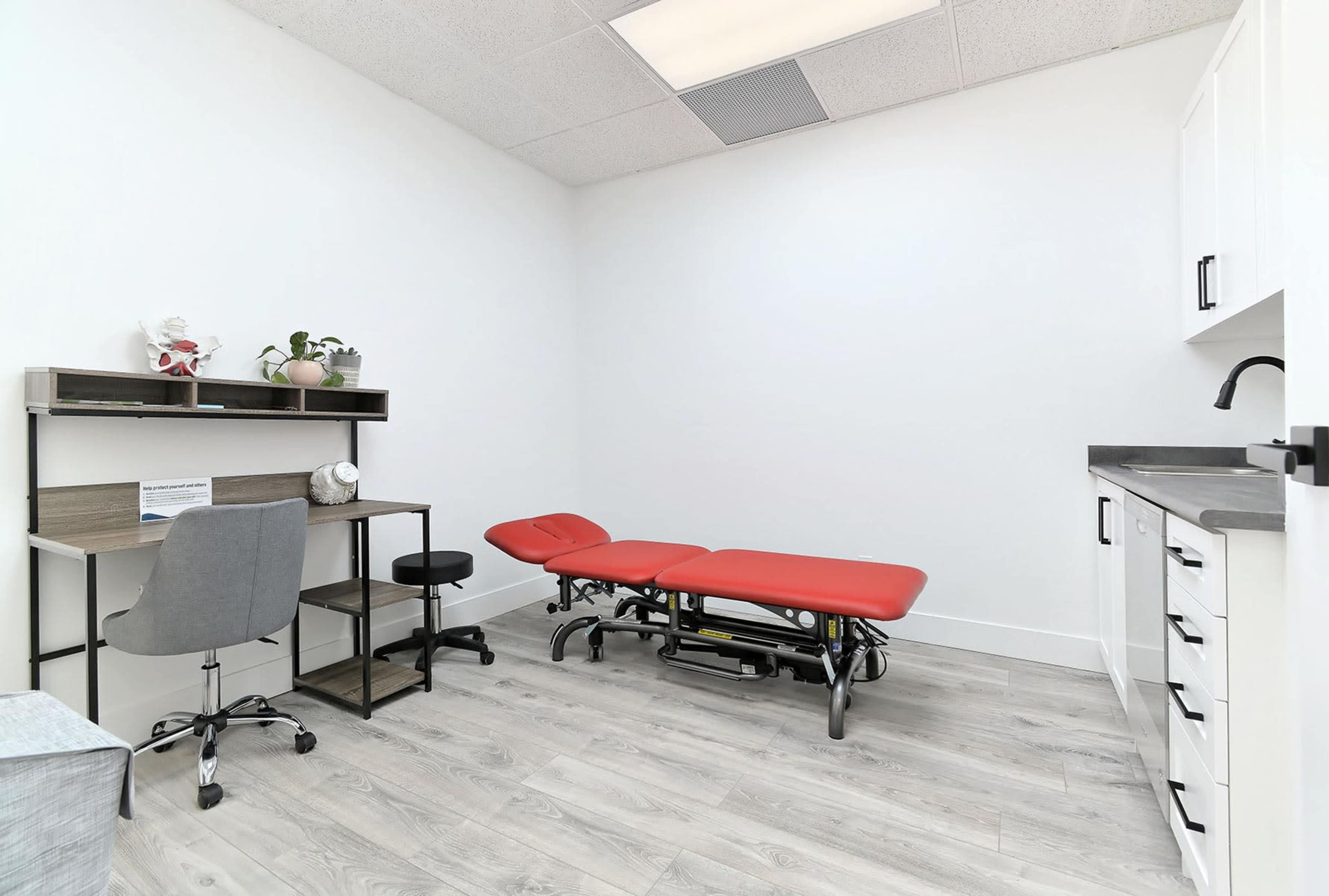 A minimalist clinical room features a red examination table, a desk with a chair, and a cabinet against a white wall.