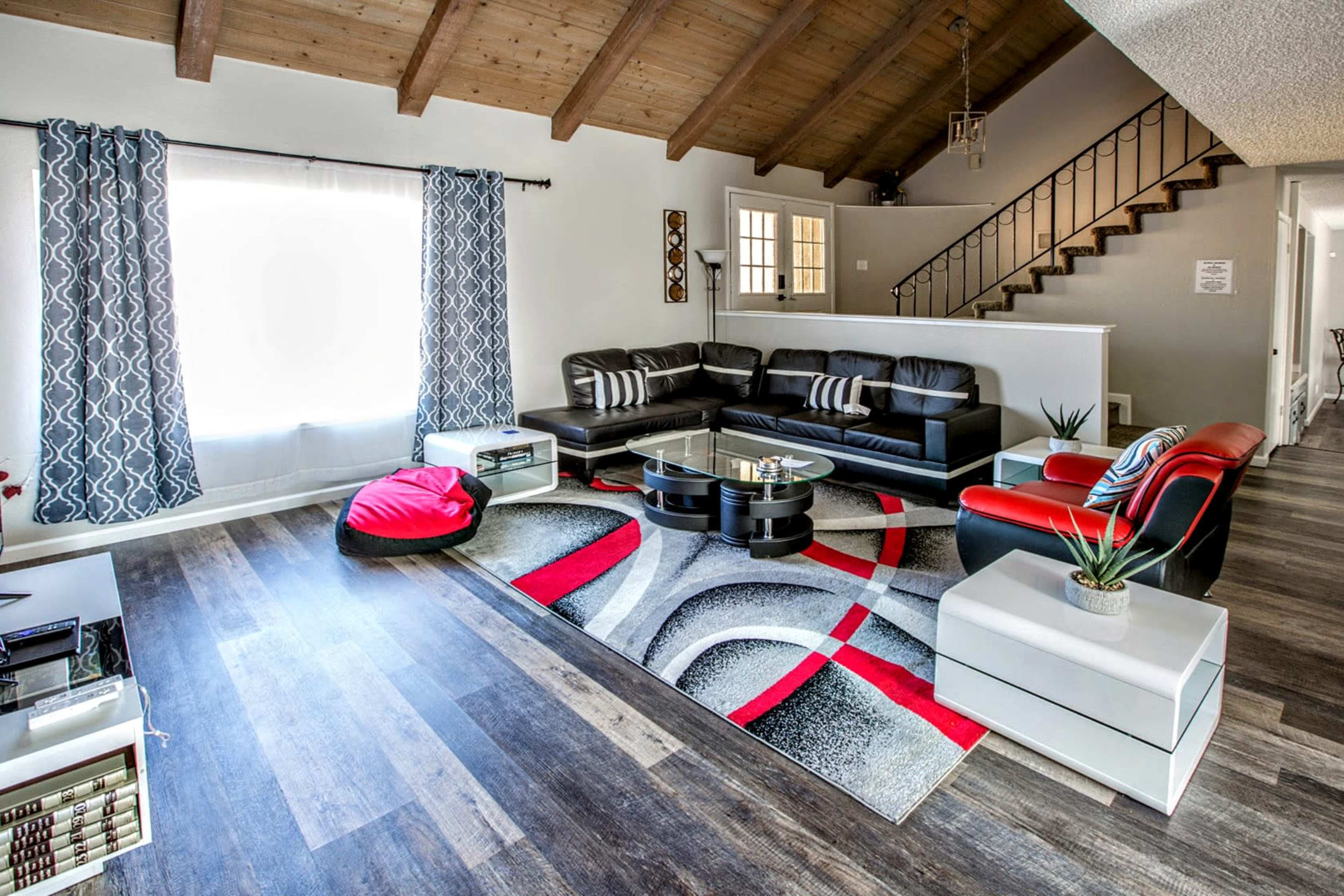 The living room features a modern black leather sectional sofa, a red accent chair, a large area rug with geometric patterns, and a high wood-beamed ceiling.