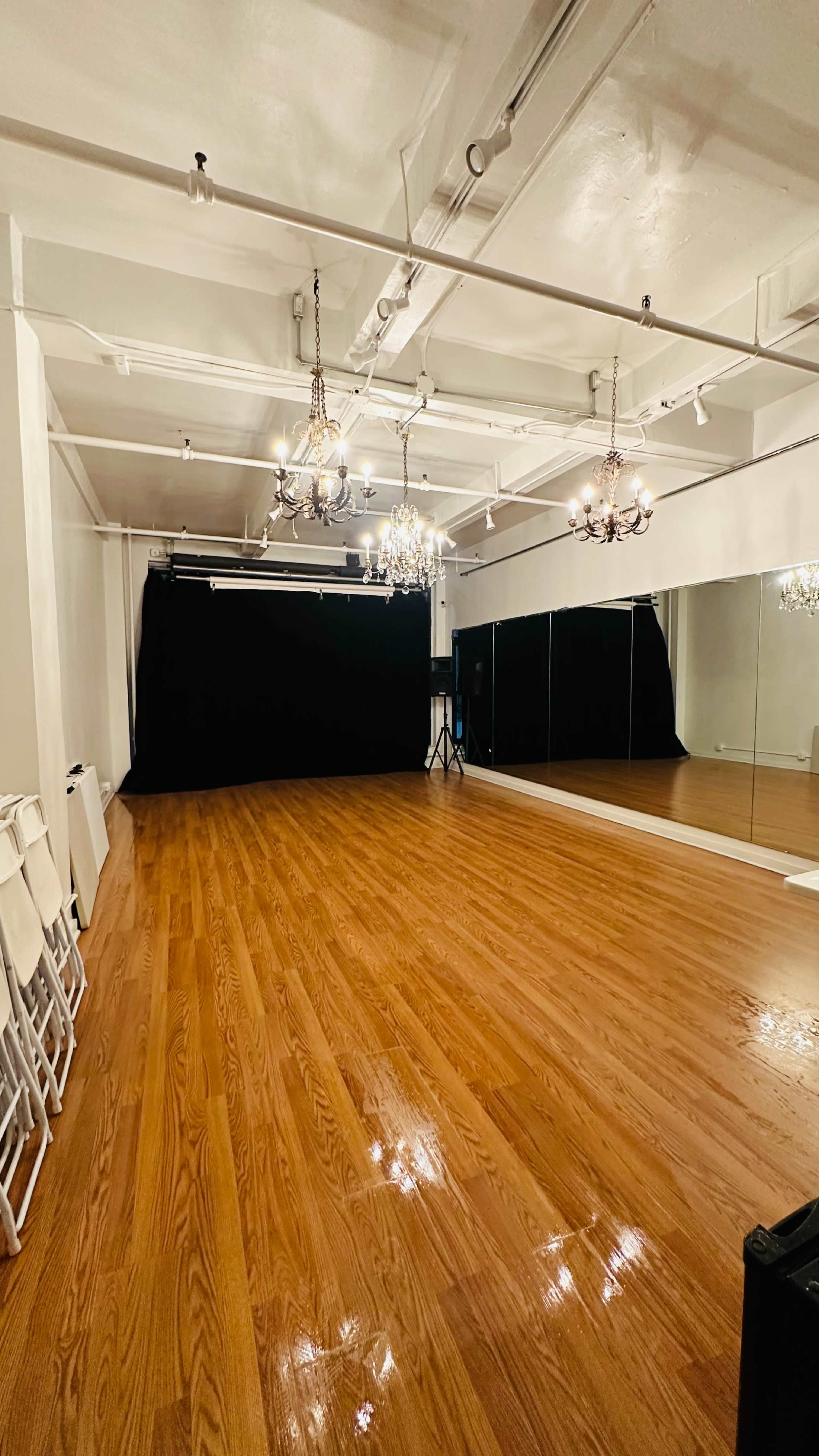 The image shows a spacious studio with polished wooden floors, a large mirror, and chandeliers hanging from the ceiling, alongside a black curtain backdrop.