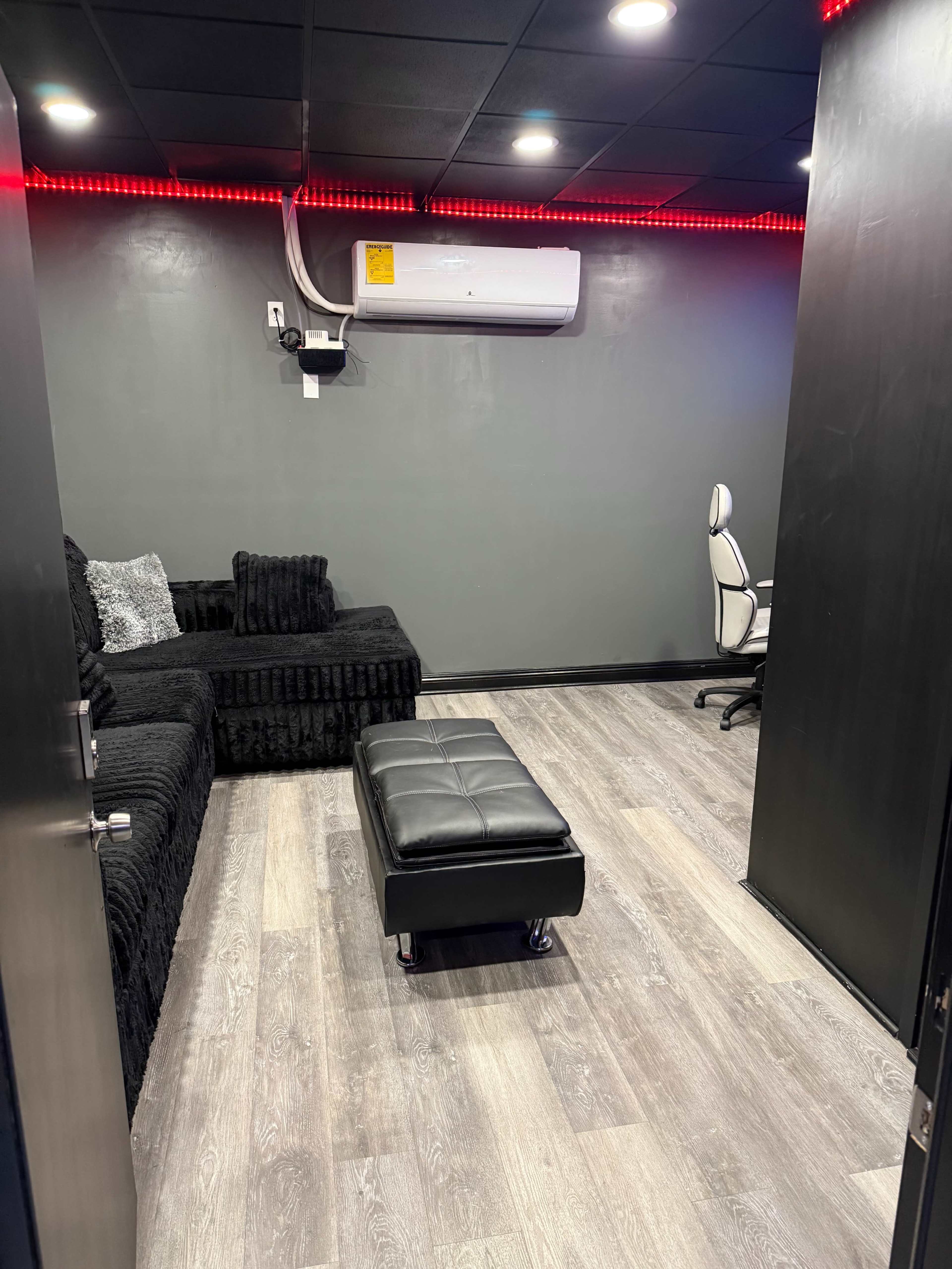 The image shows a small room featuring a black couch, a black ottoman on wheels, and a desk with a white chair, all set against a gray wall and illuminated by red LED lights.