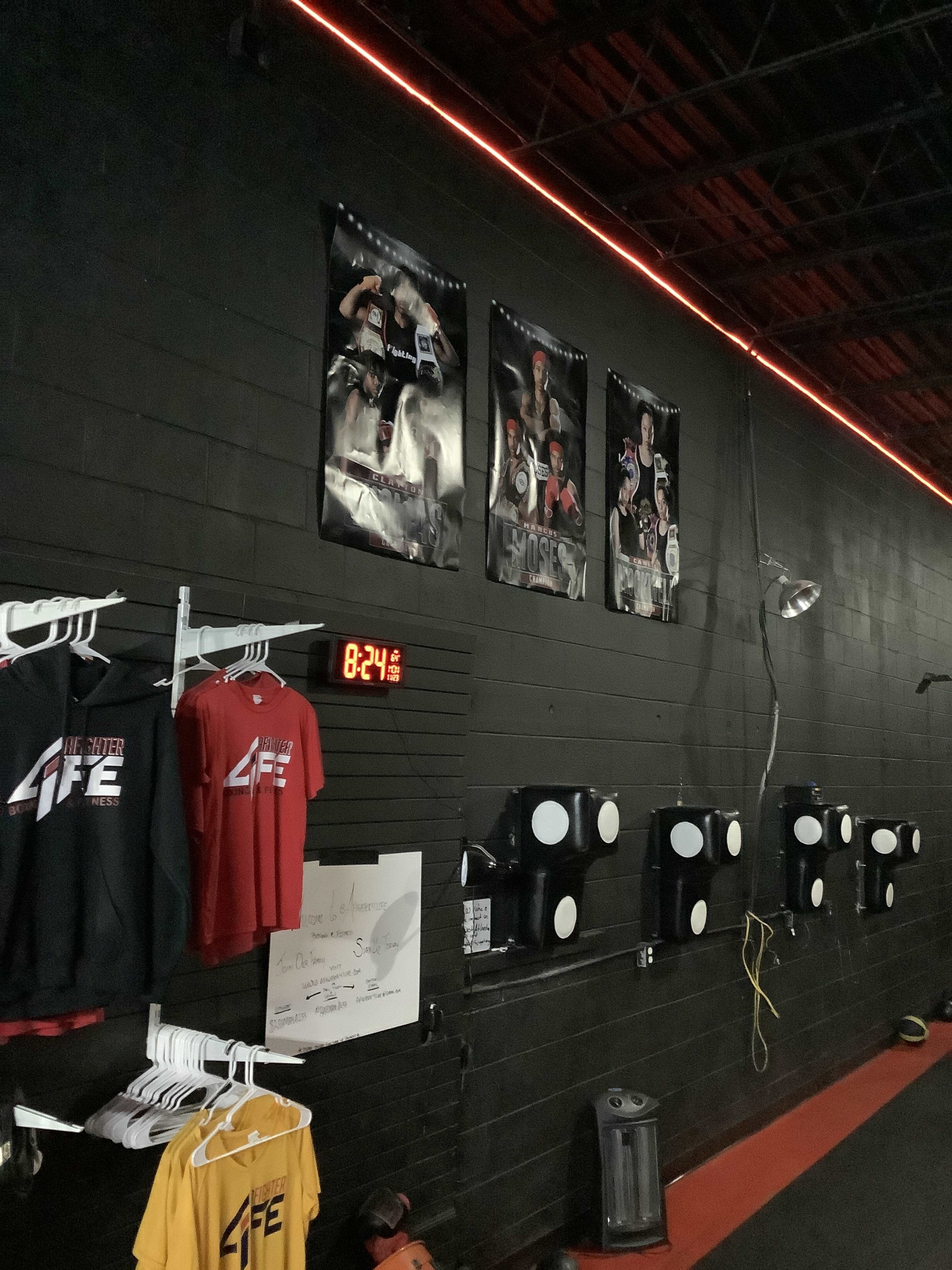 A gym wall featuring workout posters, a digital clock displaying 8:24, and a rack with red and yellow workout shirts.