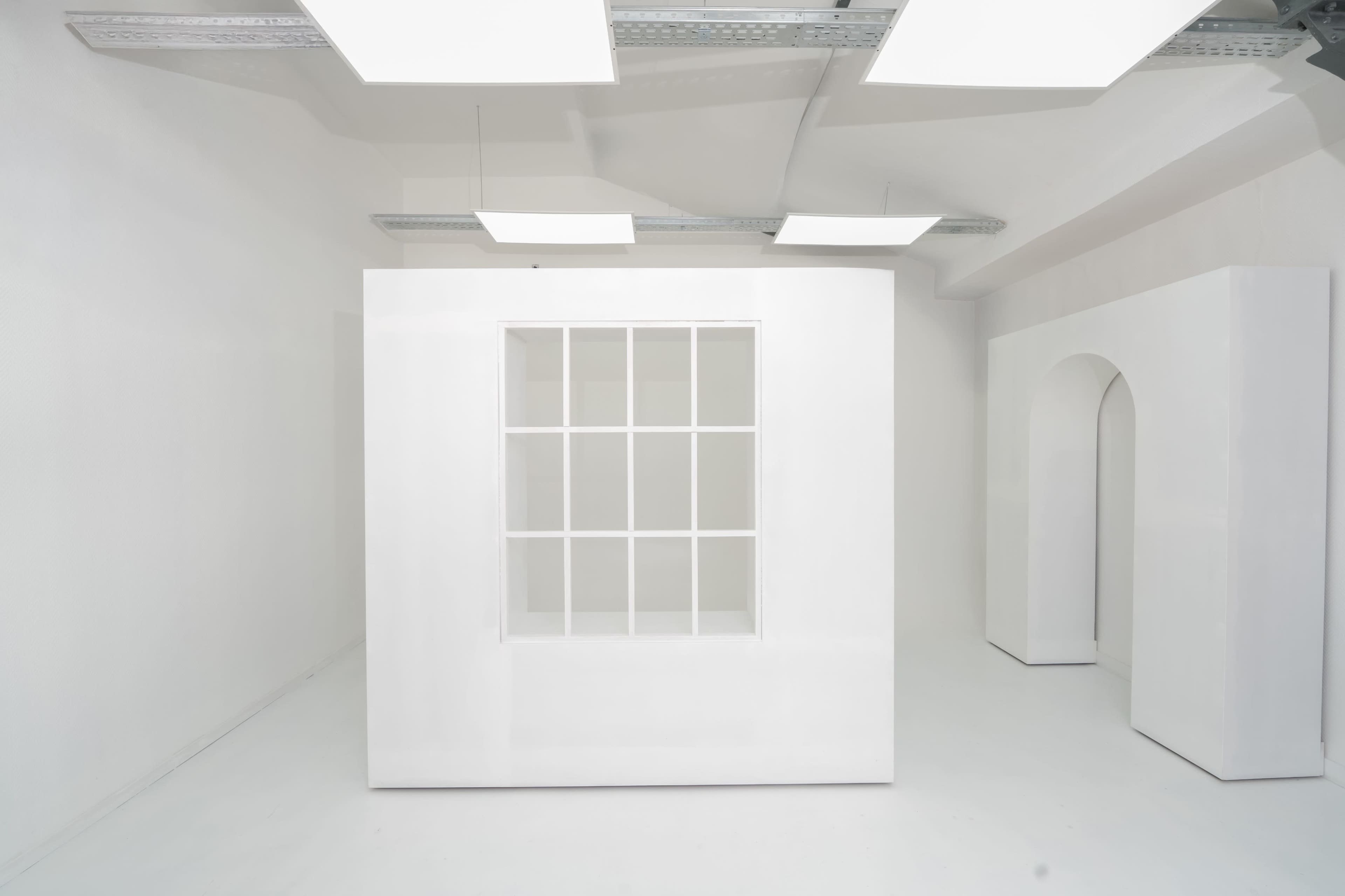 The image shows a minimalistic white room featuring a large, white box with nine square windows and a white archway on the right.