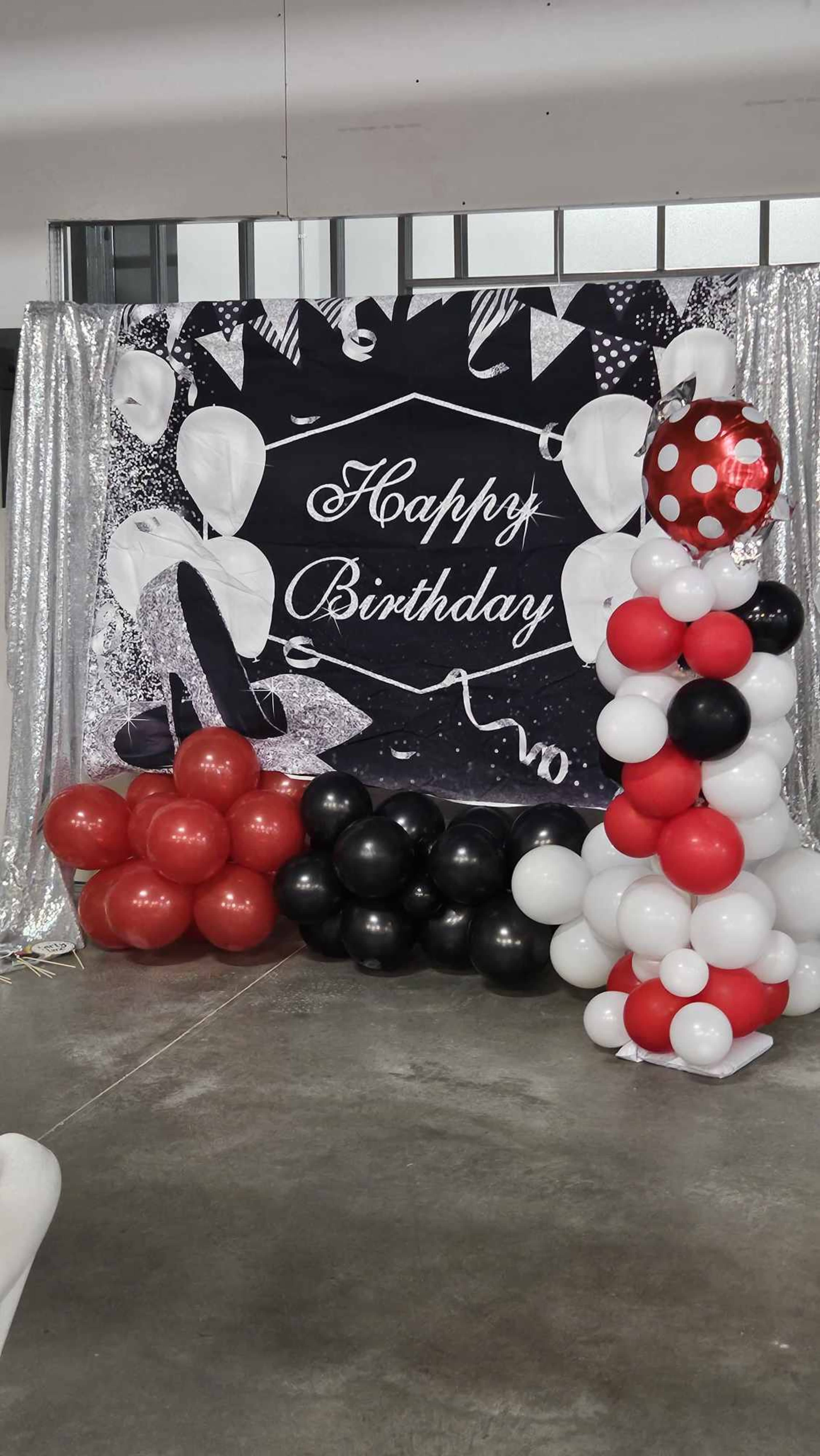 A birthday decoration featuring a backdrop with "Happy Birthday" in white text and a display of balloons in red, black, and white arranged in two clusters.