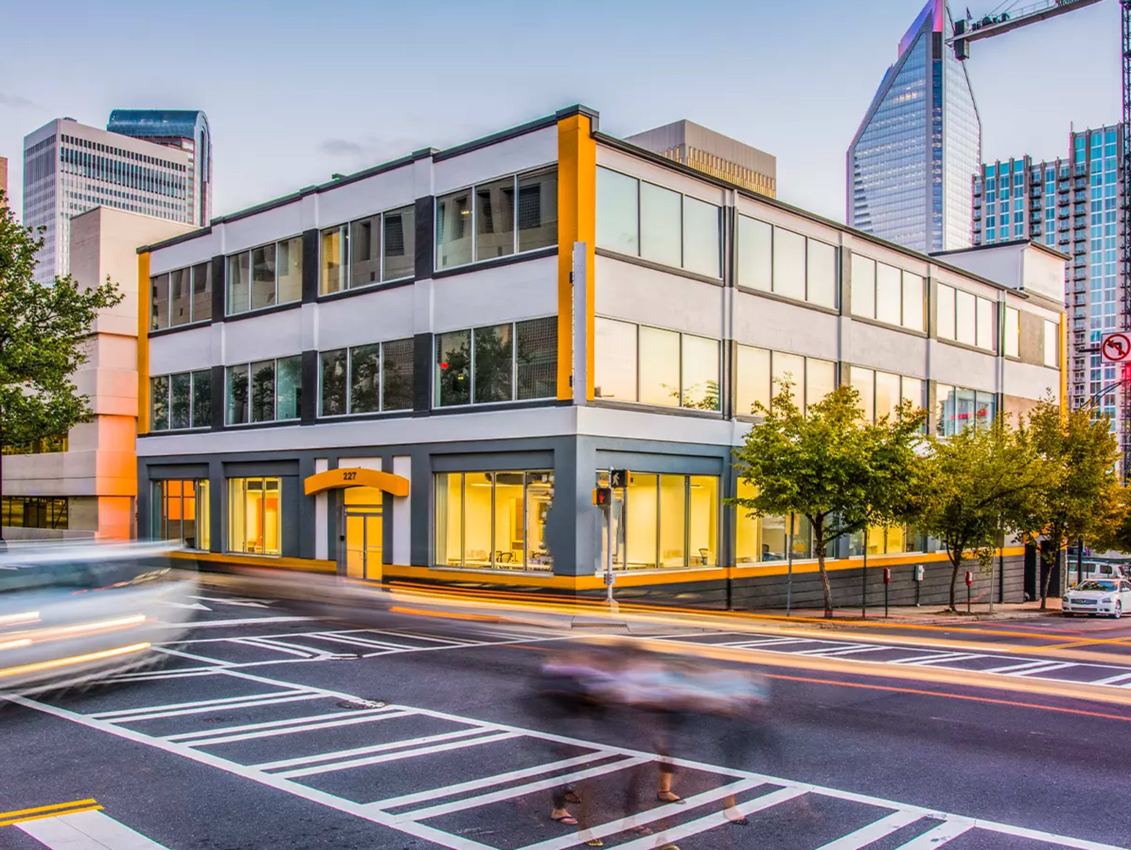 The image shows a modern three-story building at an urban intersection, with large windows and a yellow accent, while vehicles move along the road in front.