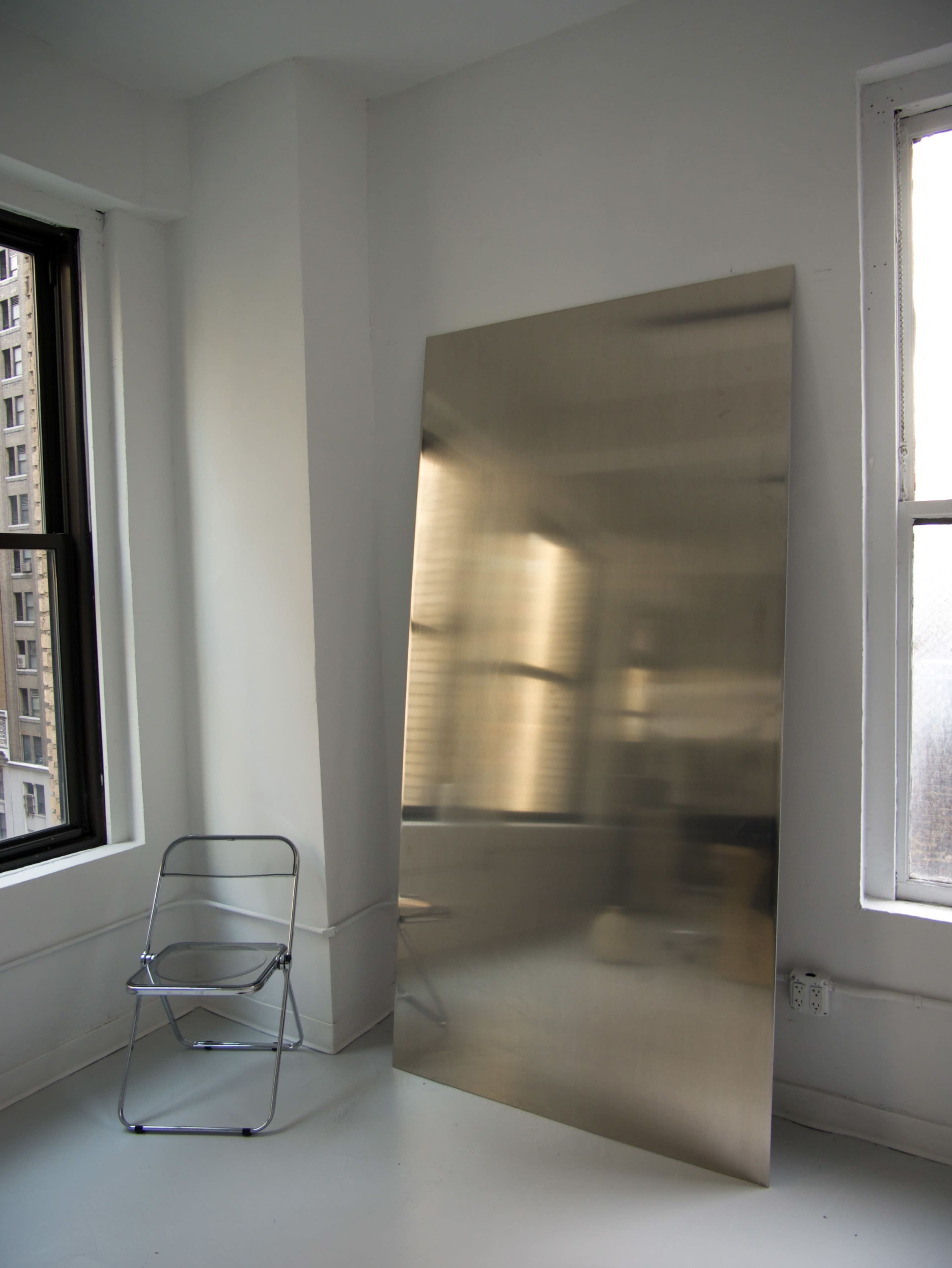 A large, reflective metal panel leans against a wall near a simple, metallic folding chair in a bright, minimalist room with windows.