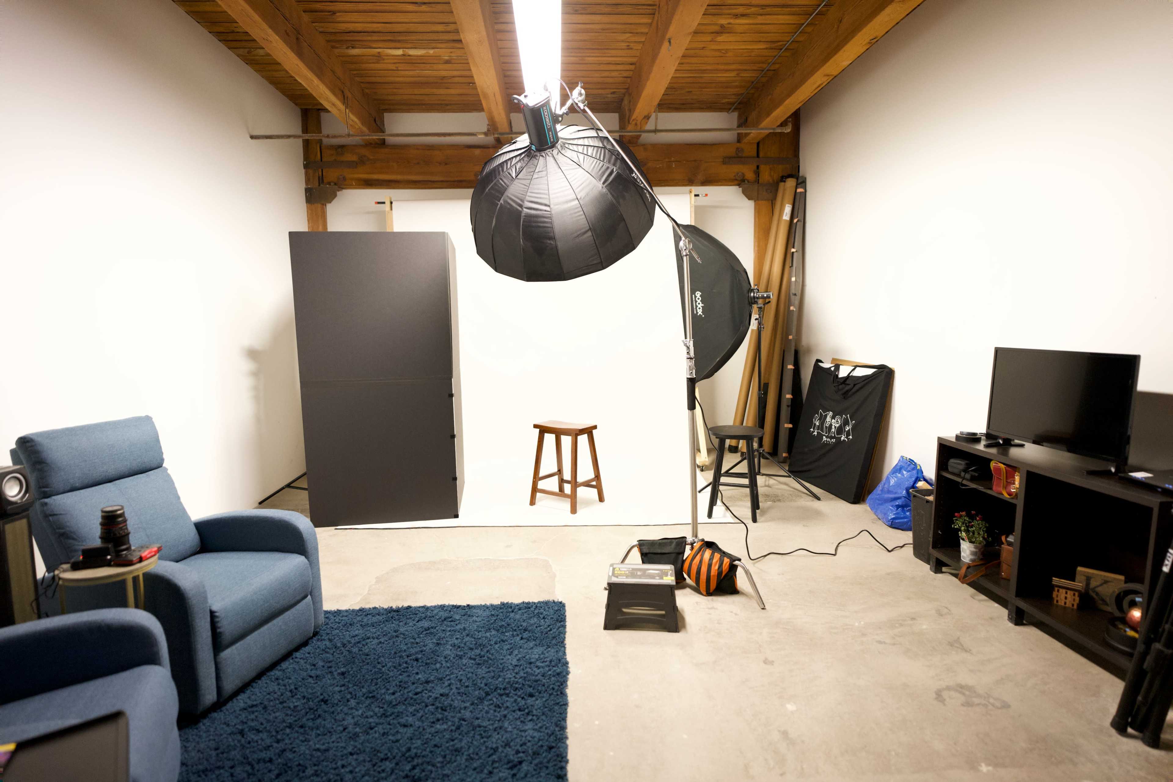 A photography studio features a blue armchair, a brown stool, lighting equipment, a TV, and a backdrop in a minimally furnished space.