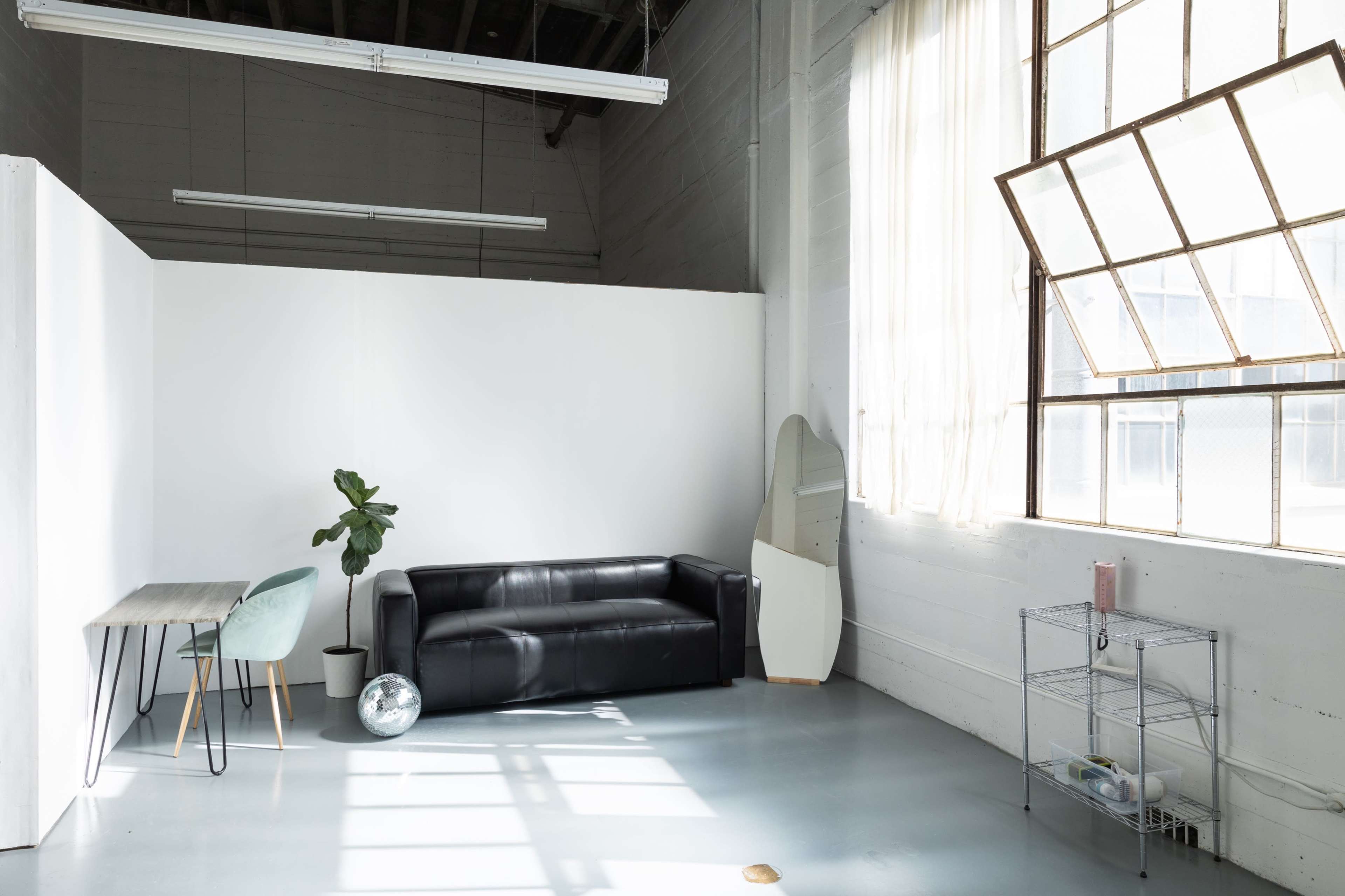 A minimalist interior features a black sofa, a light wood table with a green chair, a potted plant, and large windows that allow natural light to flow in.
