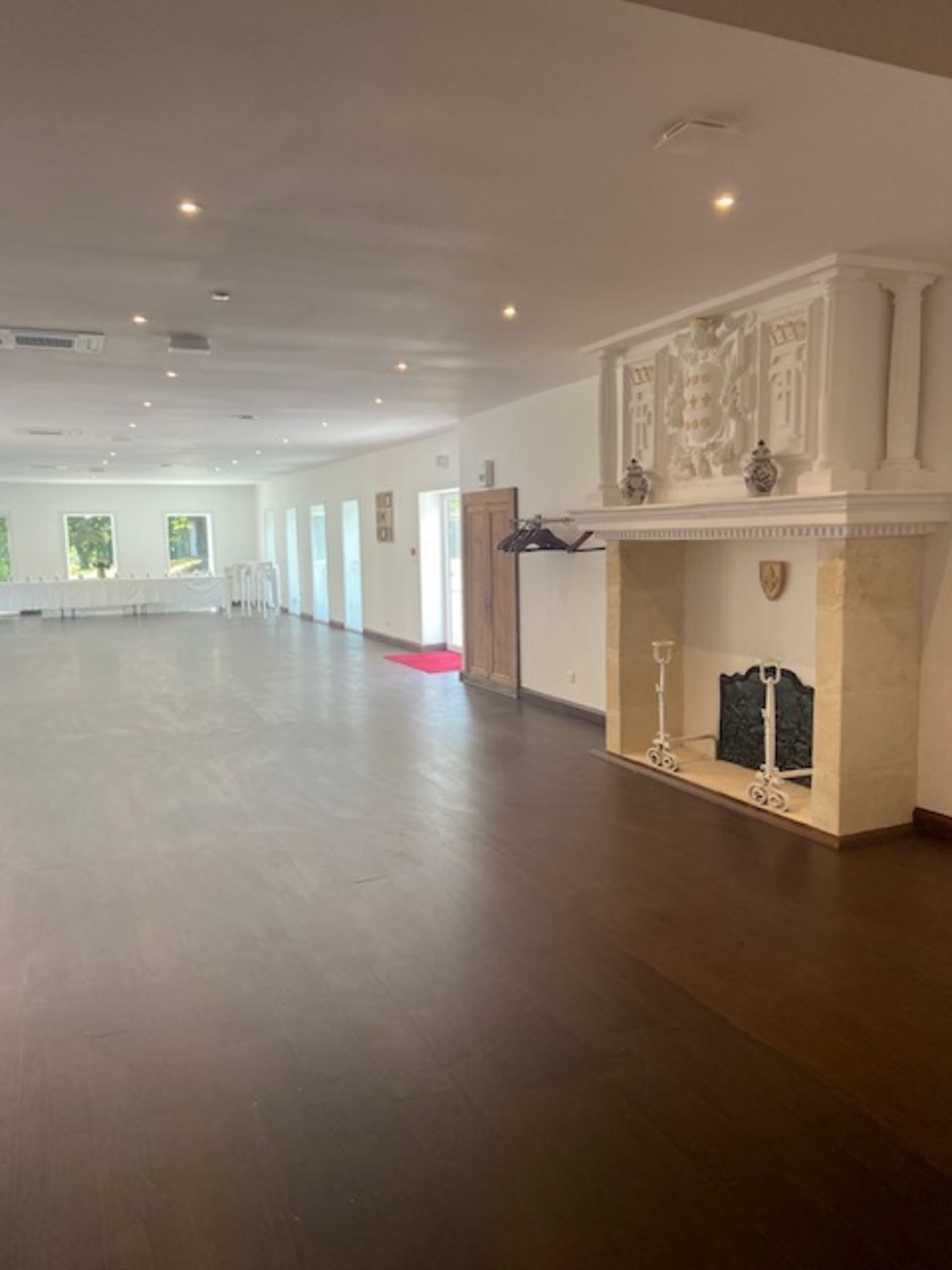 The image shows a spacious indoor room featuring a fireplace, large windows, and a table set up in the background.