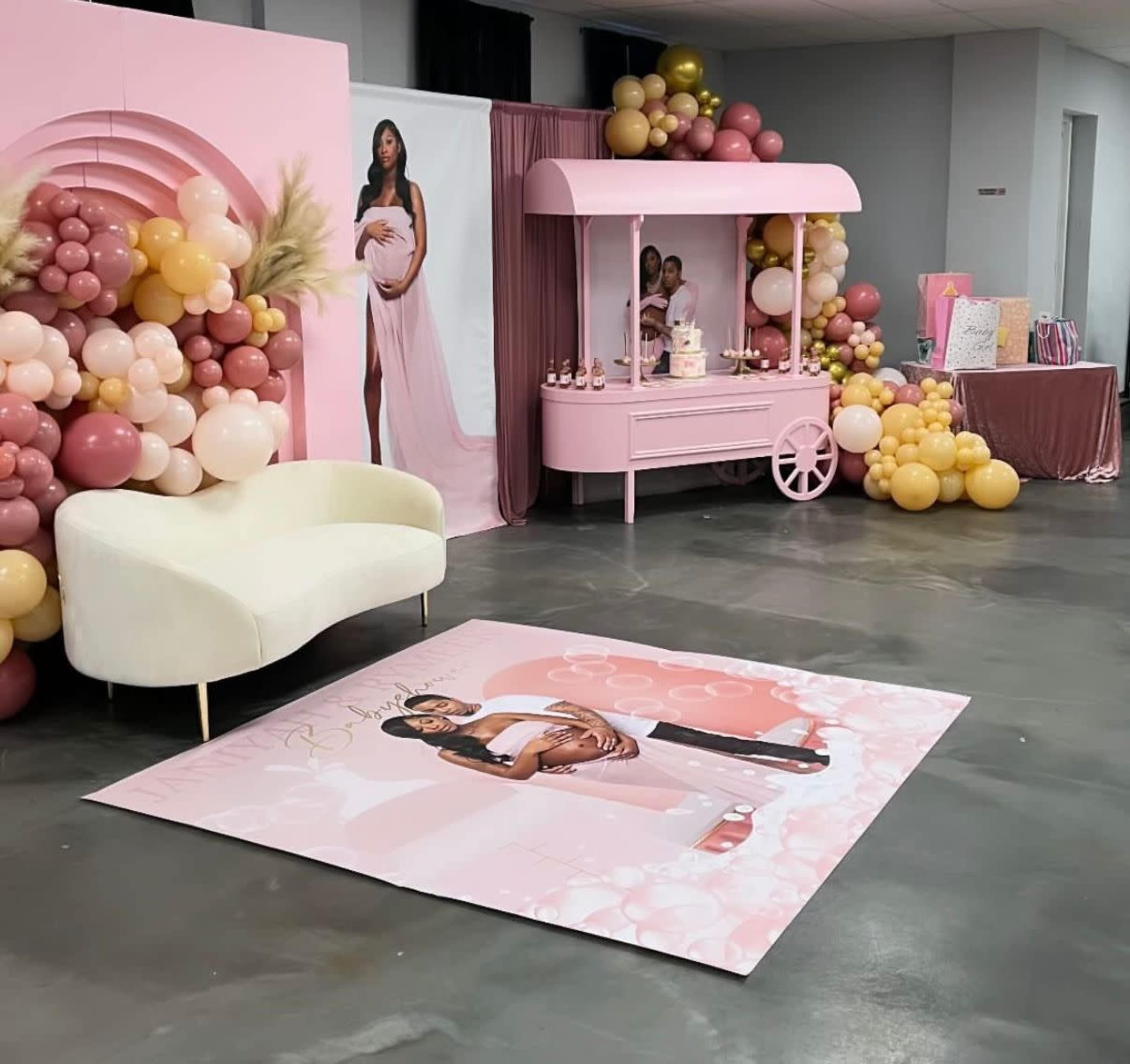 The image shows a decorated event space featuring a pink cart with a cake, a white couch, and a large backdrop adorned with balloons and a floor graphic of a couple holding a pregnant belly.