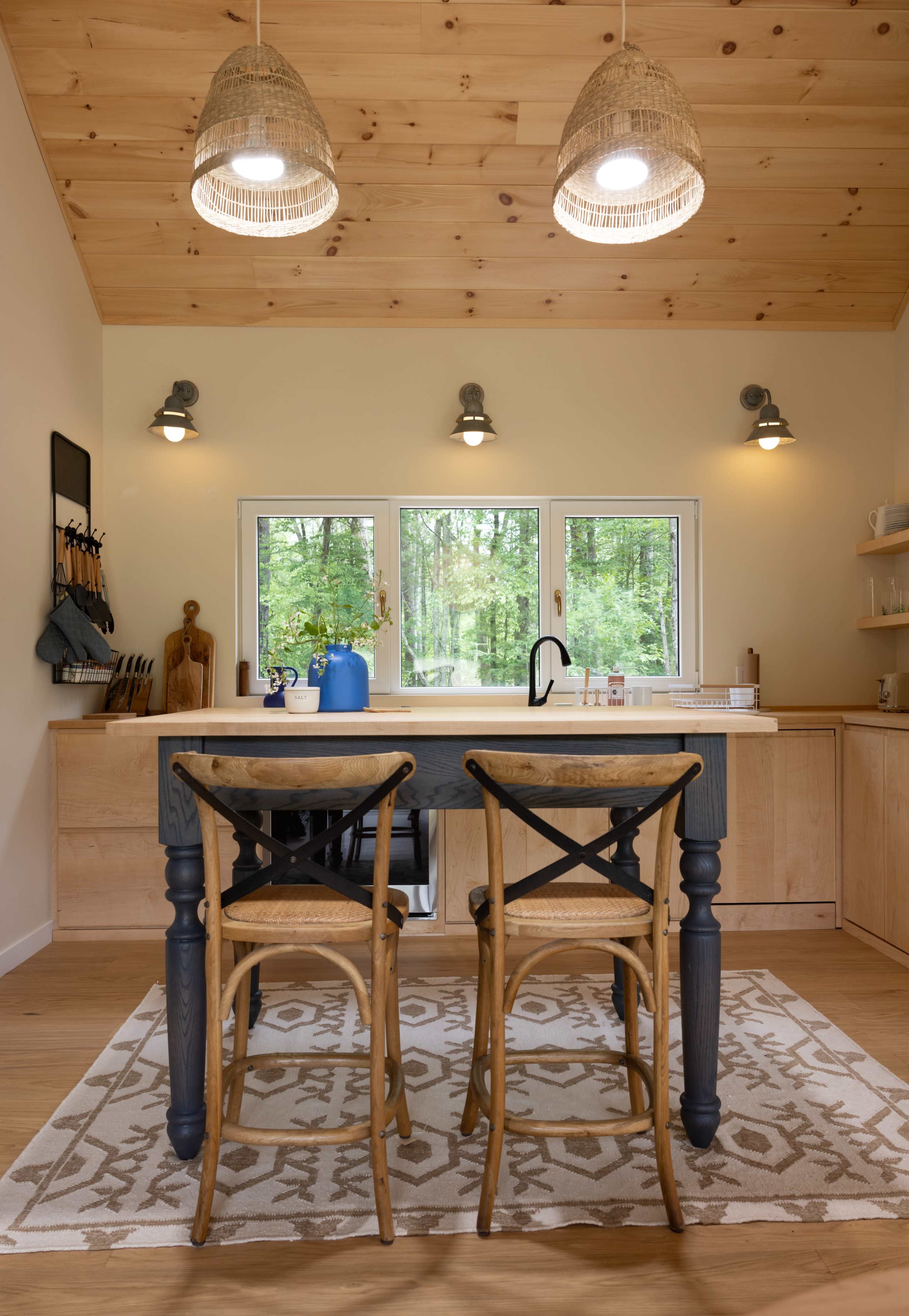 A modern kitchen features a wooden countertop with two beige bar stools, pendant lighting, and large windows overlooking a forested area.
