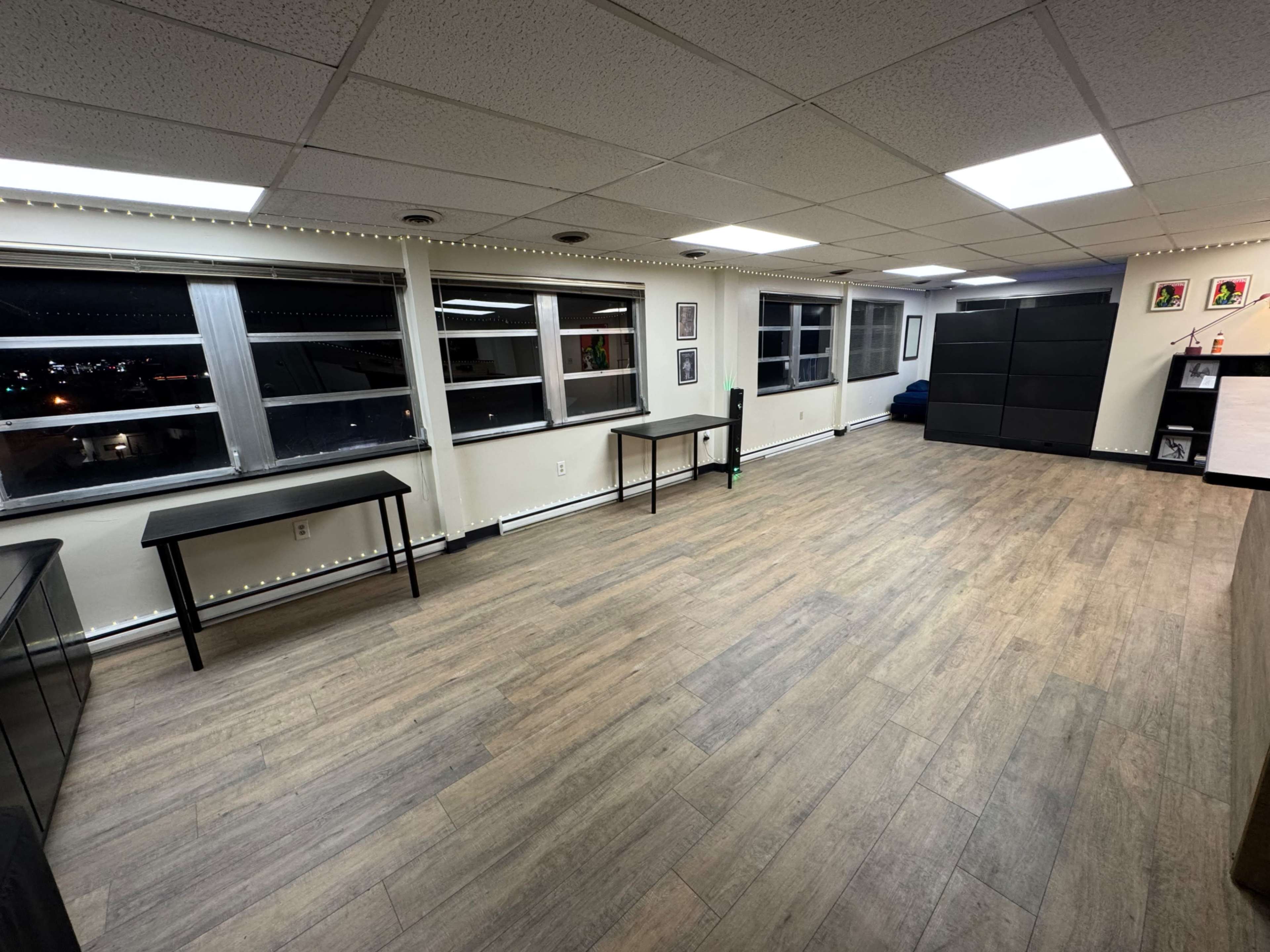 The image shows a spacious, well-lit room with wooden flooring, multiple windows, and minimal furniture, including a few tables and storage units.