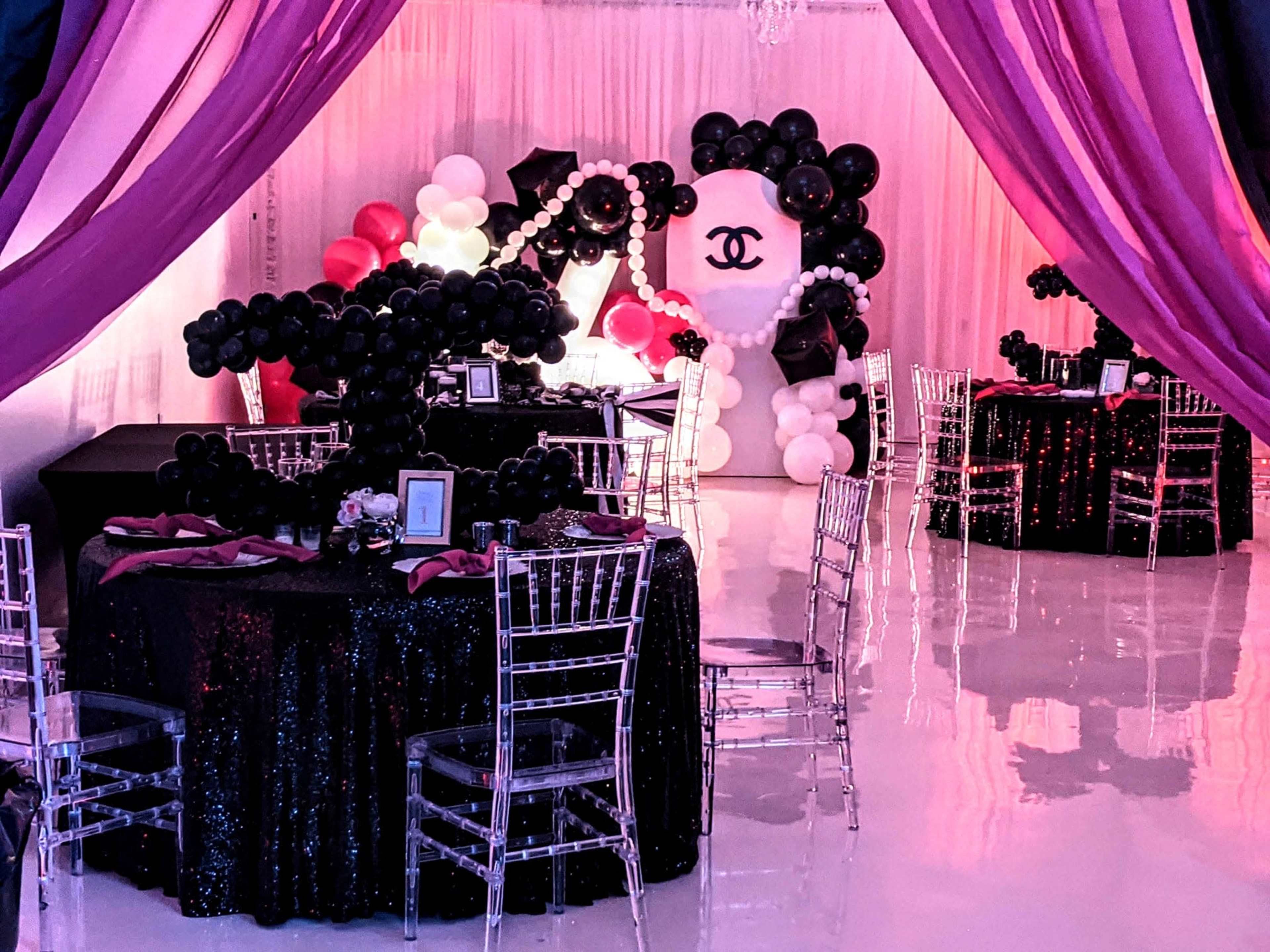 The scene features a party space adorned with black and white balloons, elegant tables with sequin tablecloths, and a backdrop shaped like a Chanel logo.