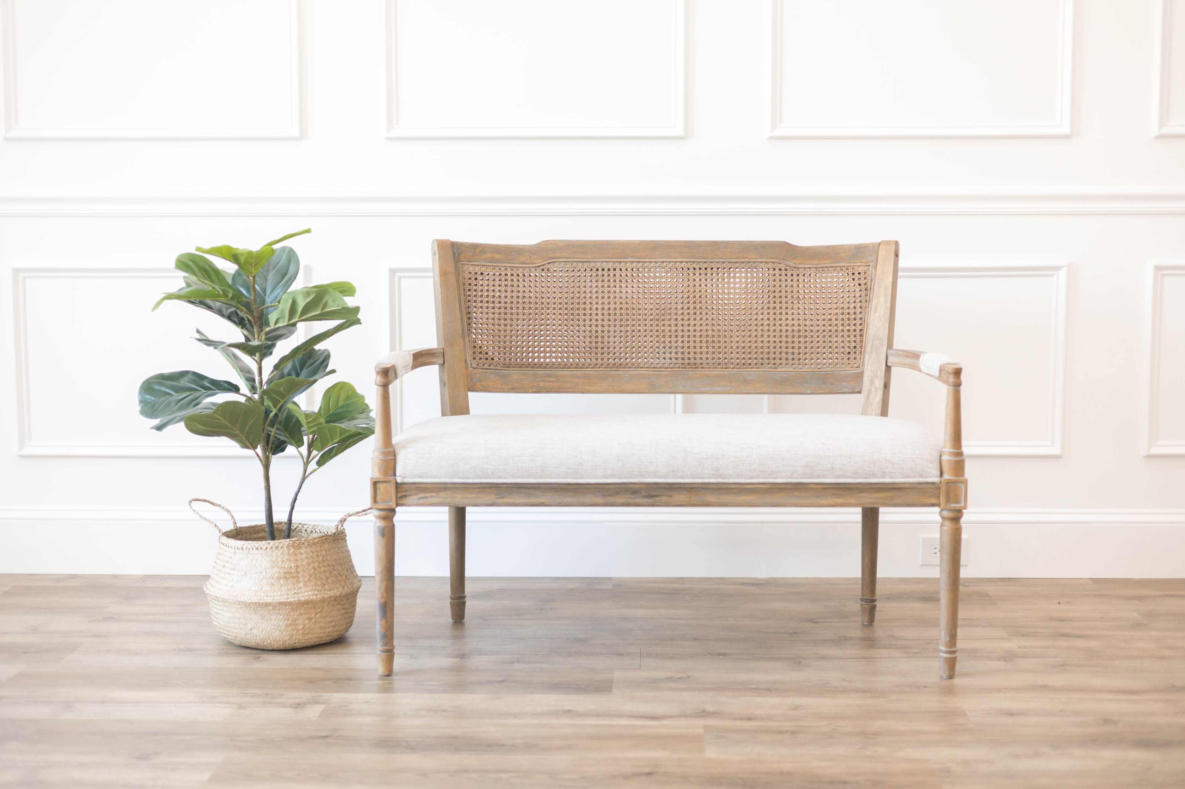 A wooden bench with a woven backrest sits beside a potted plant in a woven basket against a panelled wall.