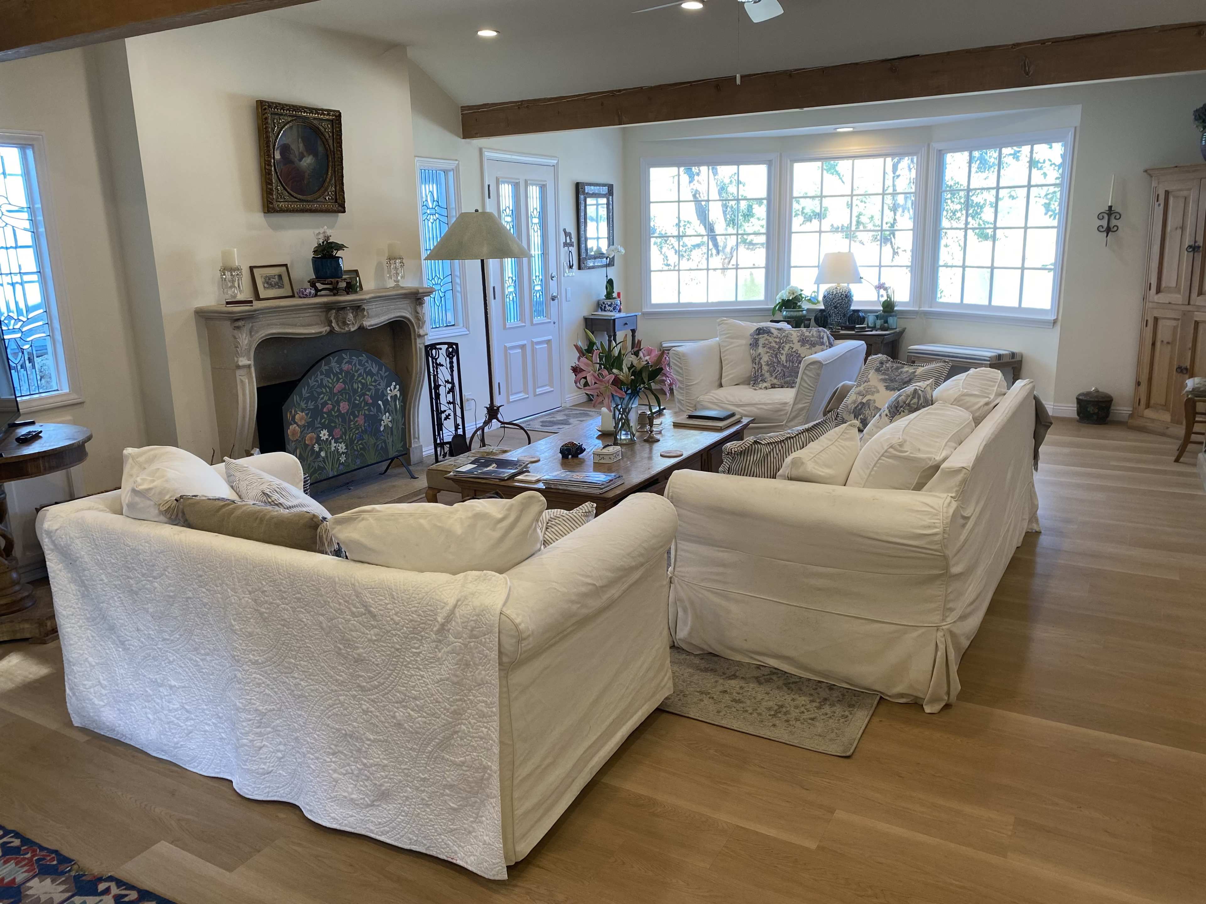The image shows a bright, open living room with white upholstered sofas arranged around a wooden coffee table, large windows, and wooden beams overhead.