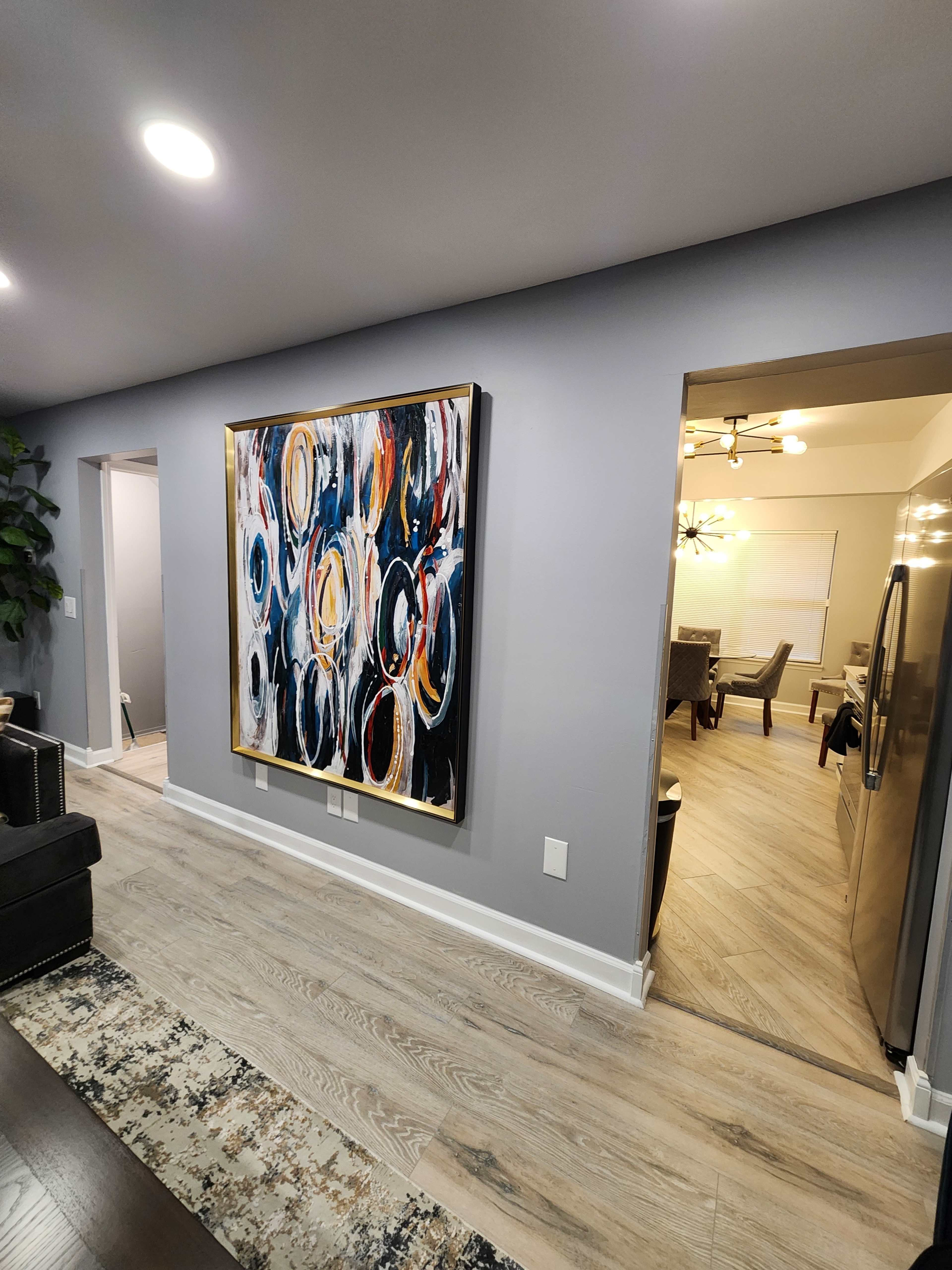 A large abstract painting with circular patterns is displayed on a gray wall, leading towards an open doorway that reveals a dining area.