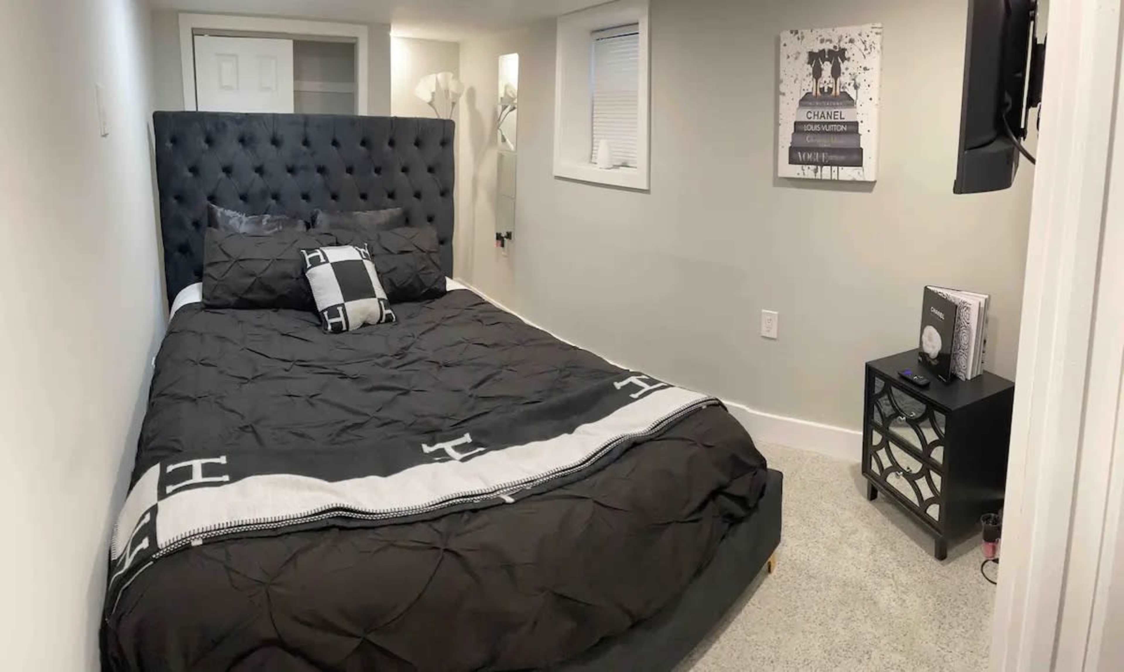 A small bedroom features a black tufted bed with a decorative blanket, a nightstand, and a wall-mounted TV.