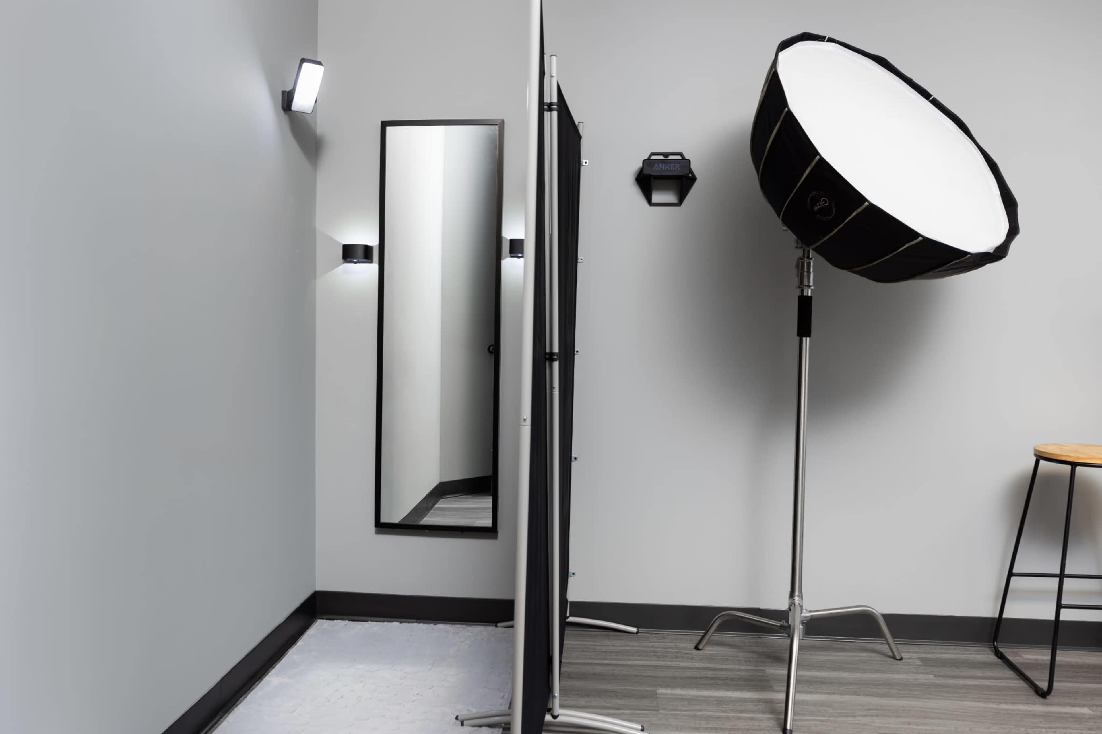The image shows a minimalist photography studio featuring a large softbox light, a mirror, a folding screen, and a stool against a gray wall.