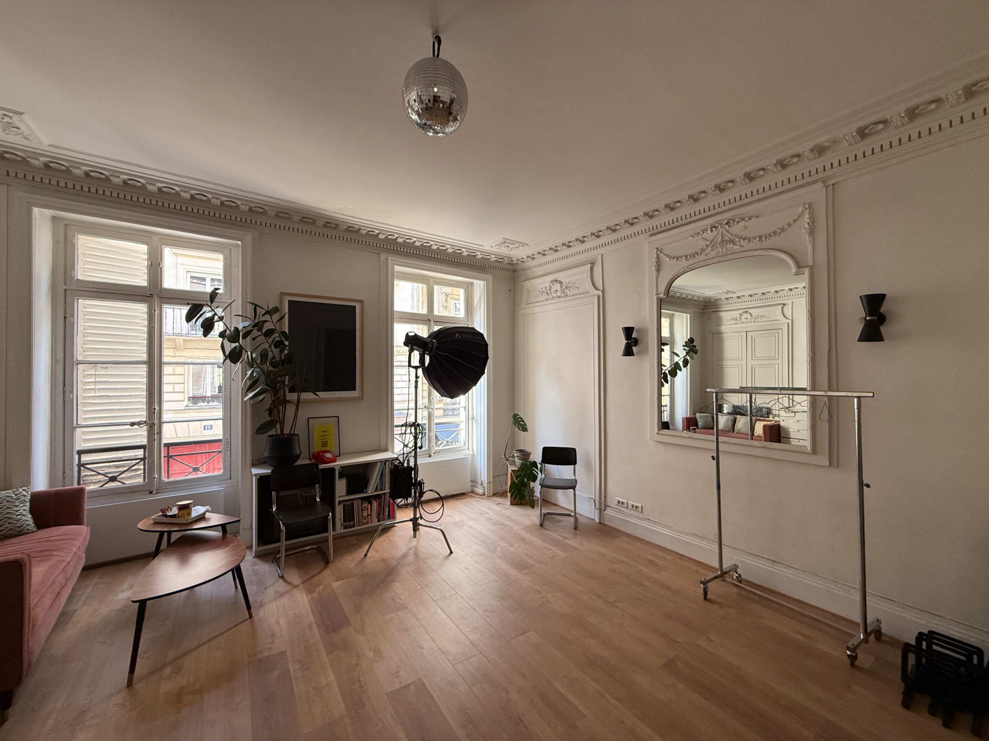 The image shows a spacious living room featuring a pink sofa, large windows, a mirror, and a floor lamp, with wooden flooring throughout.