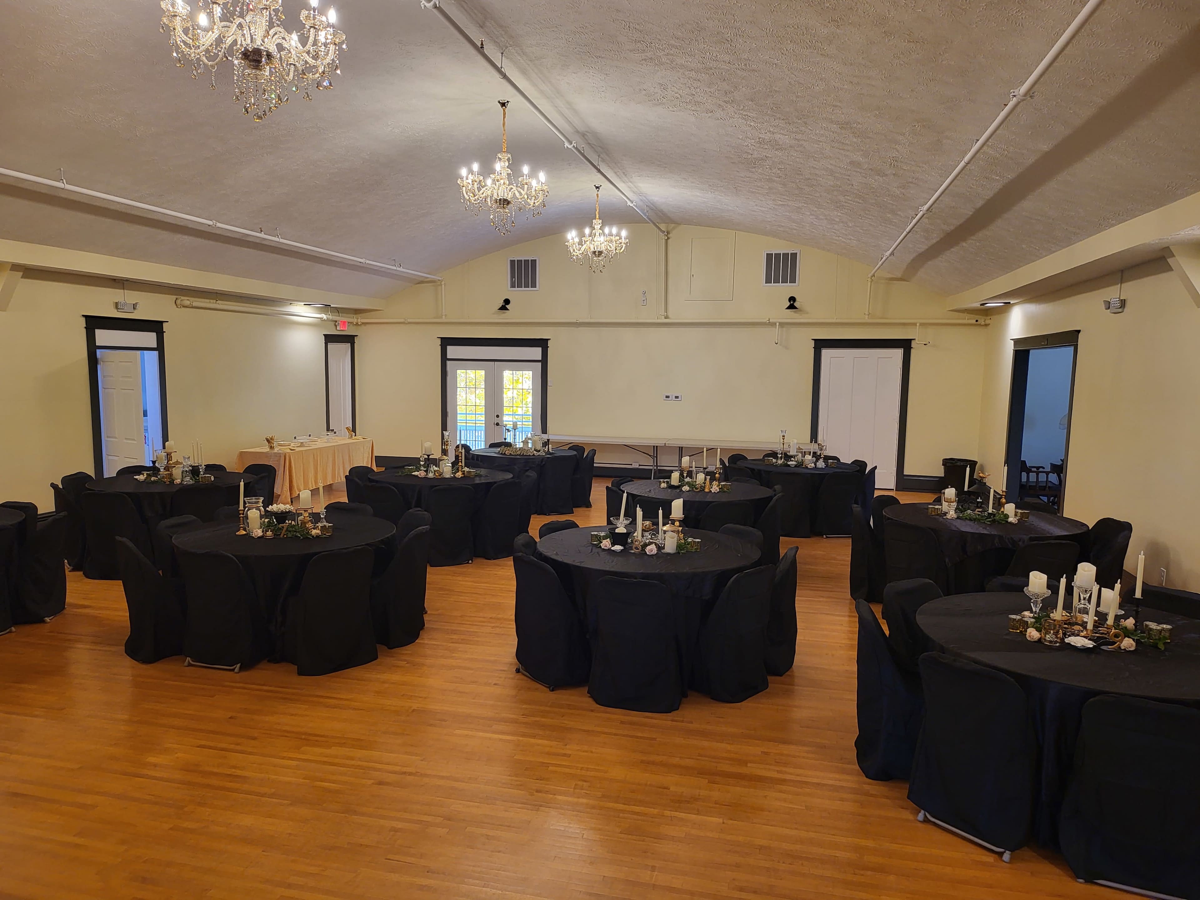 A large event space features several round tables covered in black tablecloths, each adorned with centerpieces, under a ceiling with chandeliers.