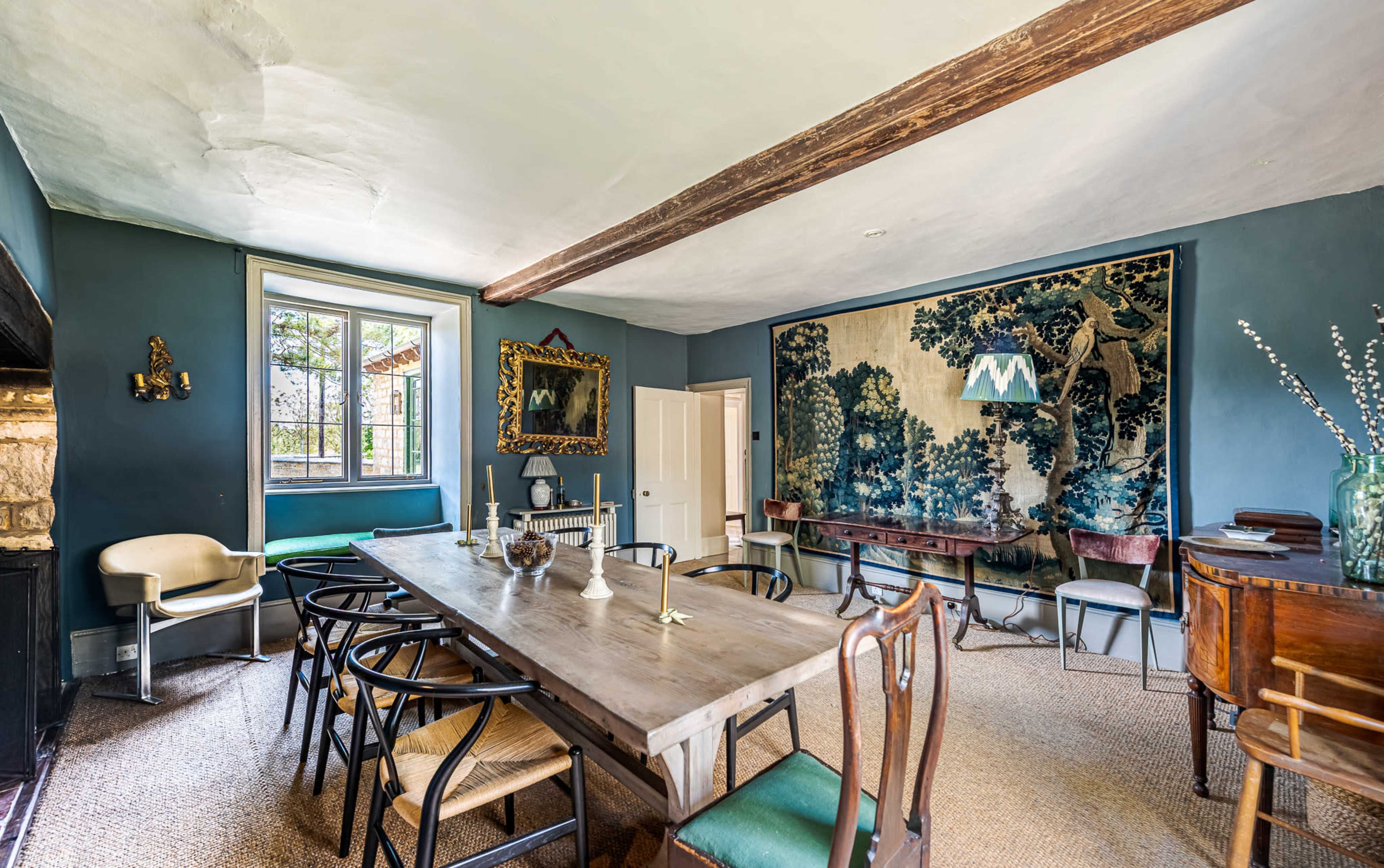 The image shows a dining room with a long wooden table, various chairs, a tapestry on the wall, and a large window that provides natural light.