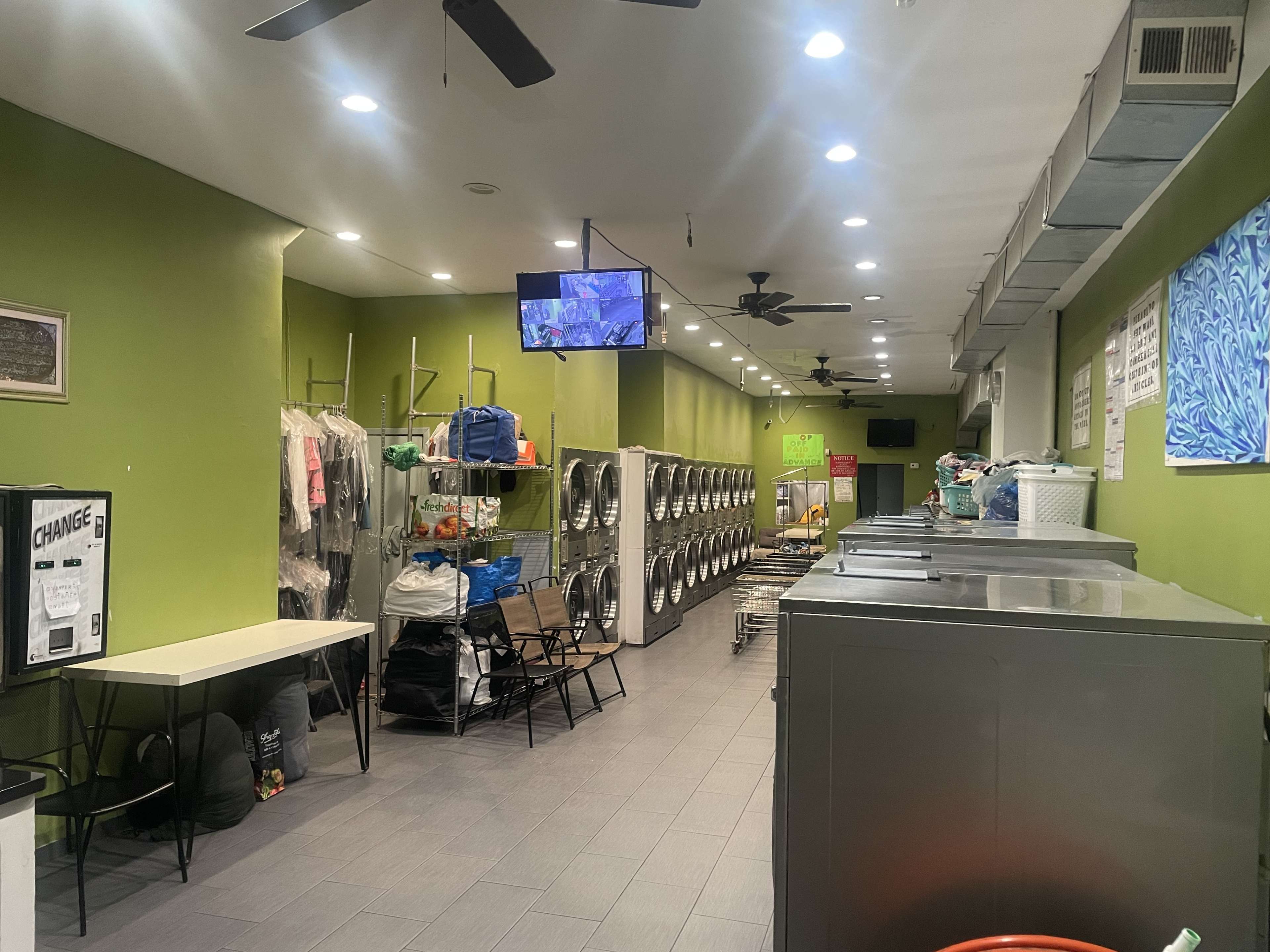 A laundry room with washing machines and dryers lined up against green walls, accompanied by folding tables and a television mounted on the wall.