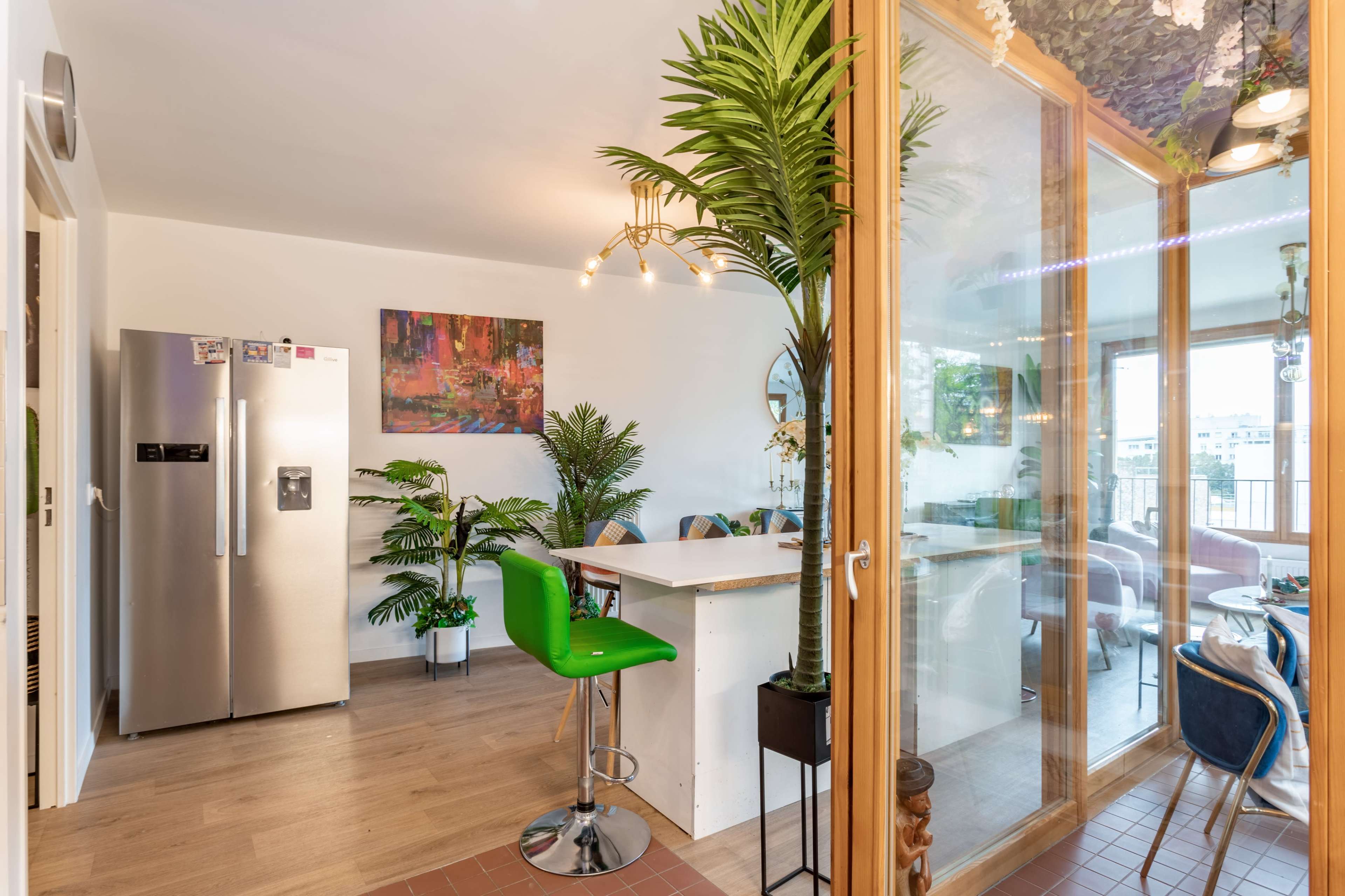 A modern kitchen area features a glass-walled dining space with a tall green bar stool, a stainless steel refrigerator, and indoor plants.