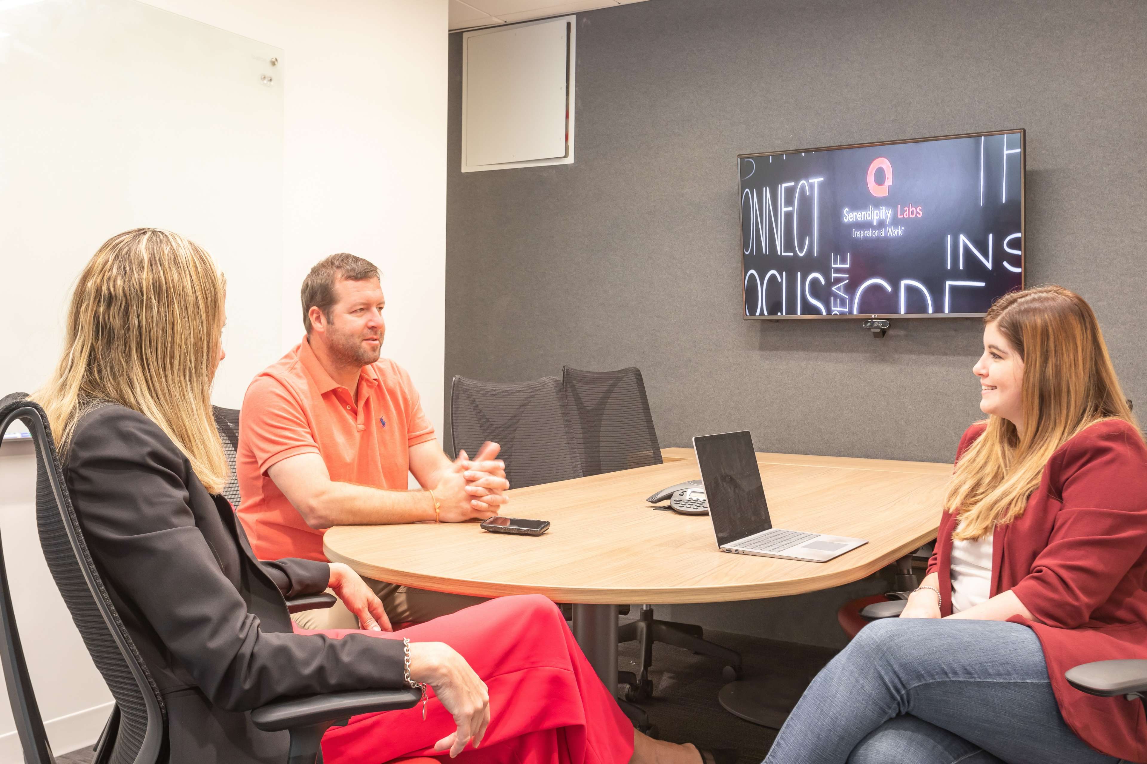 Three individuals are seated around a round conference table in a modern meeting room, discussing while a laptop and smartphone are placed on the table and a screen displays a logo in the background.