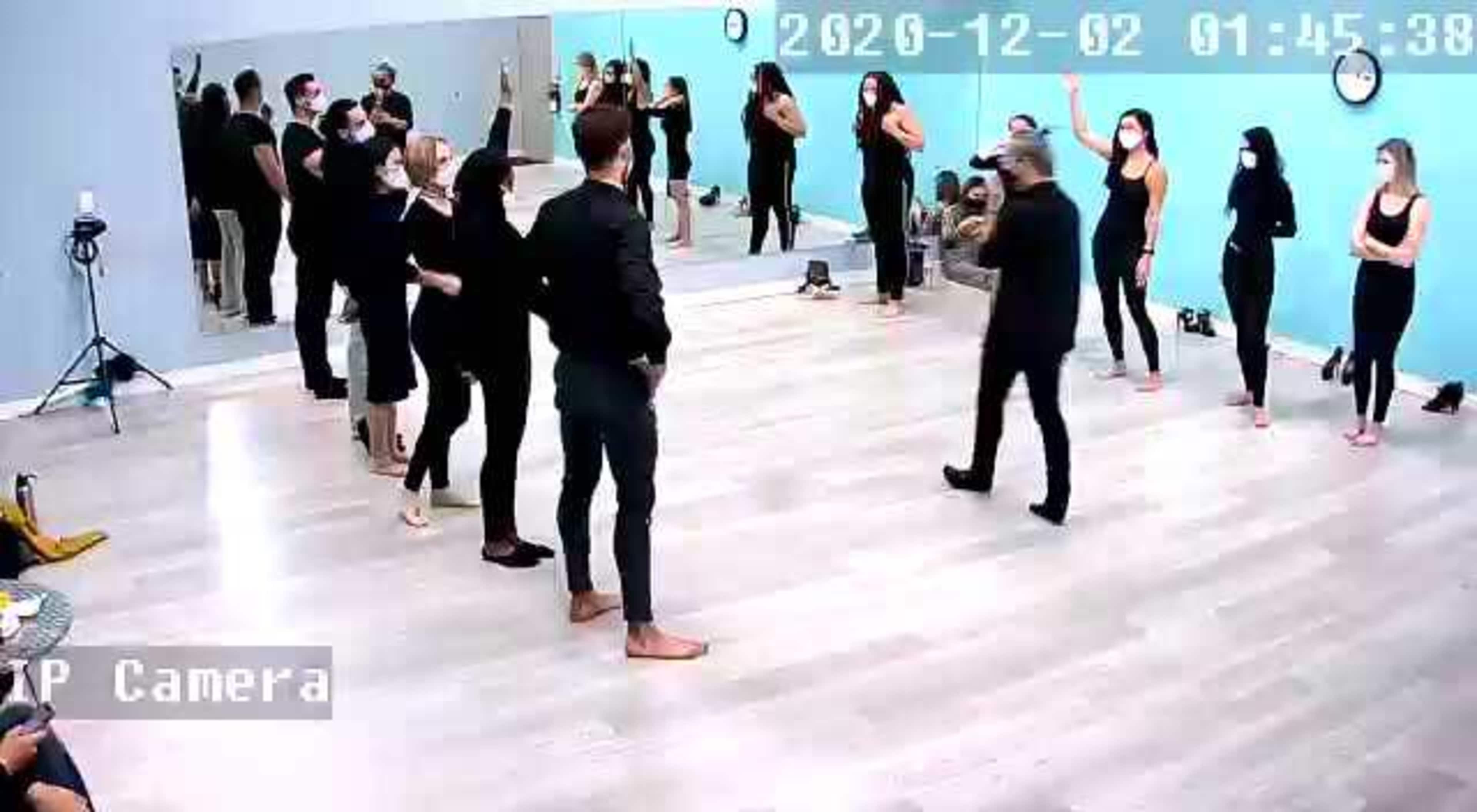 A group of people in black attire is gathered in a dance studio, engaging in a rehearsal, while some wear masks and others stand in various poses.