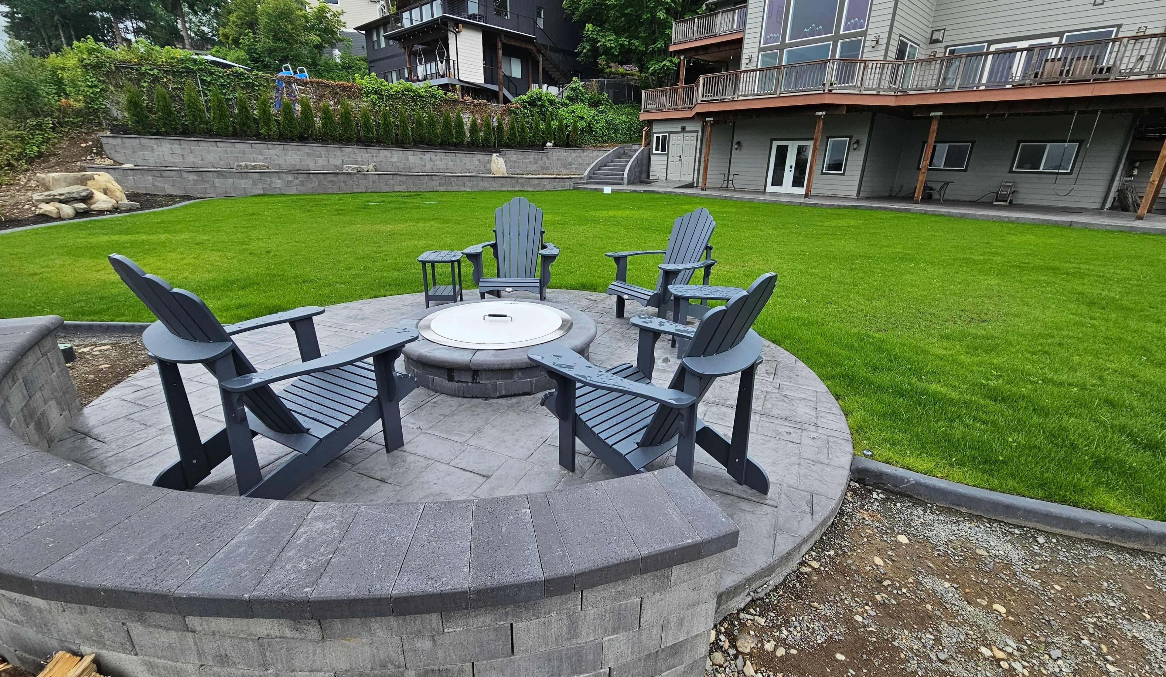 The image shows a circular fire pit area surrounded by six gray Adirondack chairs, set on a grassy lawn with landscaped stone features.