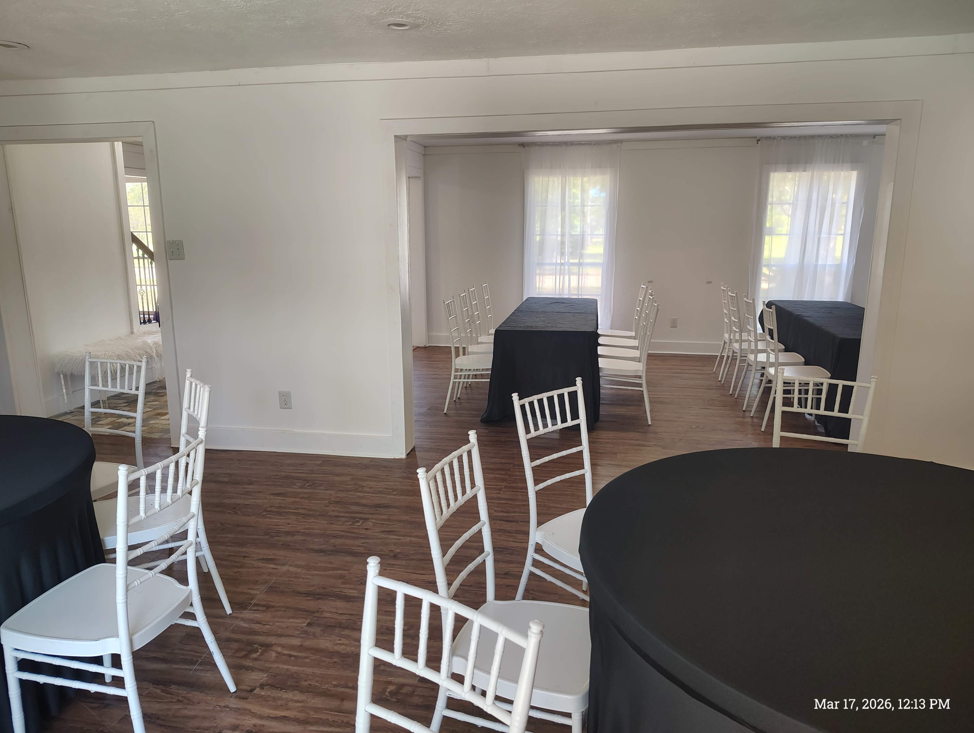 The image shows a spacious event room with several tables covered in black cloth and white chairs arranged around them, illuminated by natural light from large windows.